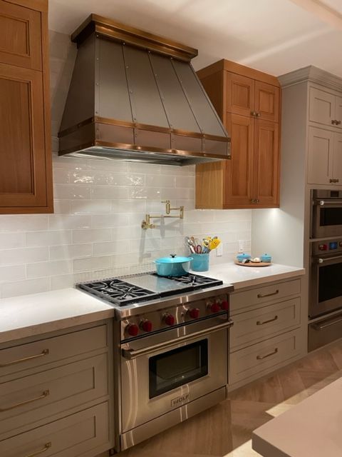 Modern kitchen with stainless steel stove, bronze range hood, white backsplash, and wood cabinets