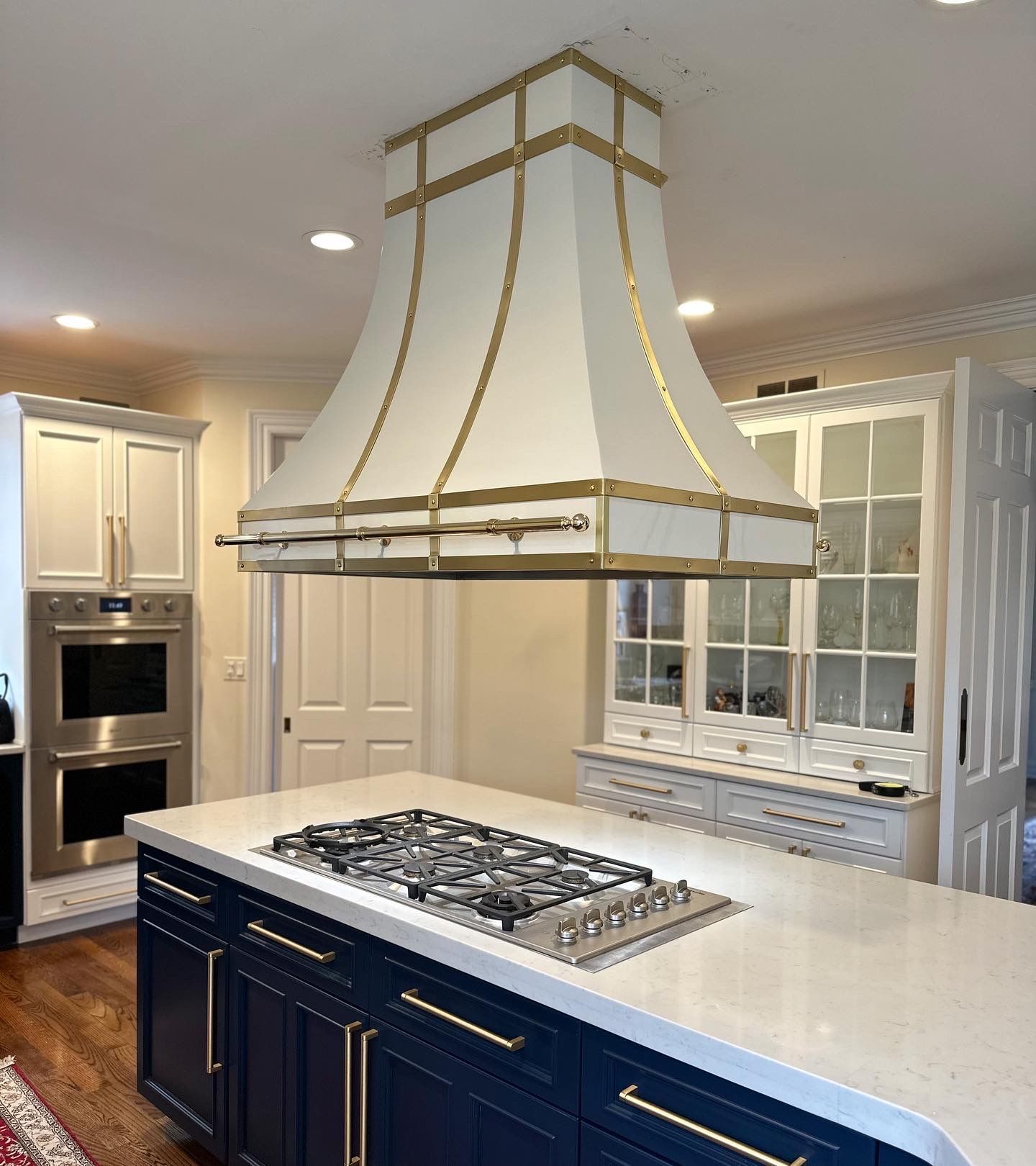 Modern kitchen with large island, white cabinetry, built-in appliances, and ornate gold-trimmed range hood over stove.