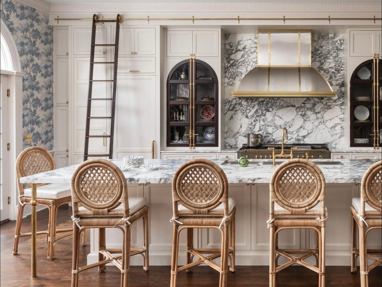 Bright white kitchen with wicker bar stools, marble island, and arched shelving with gold accents