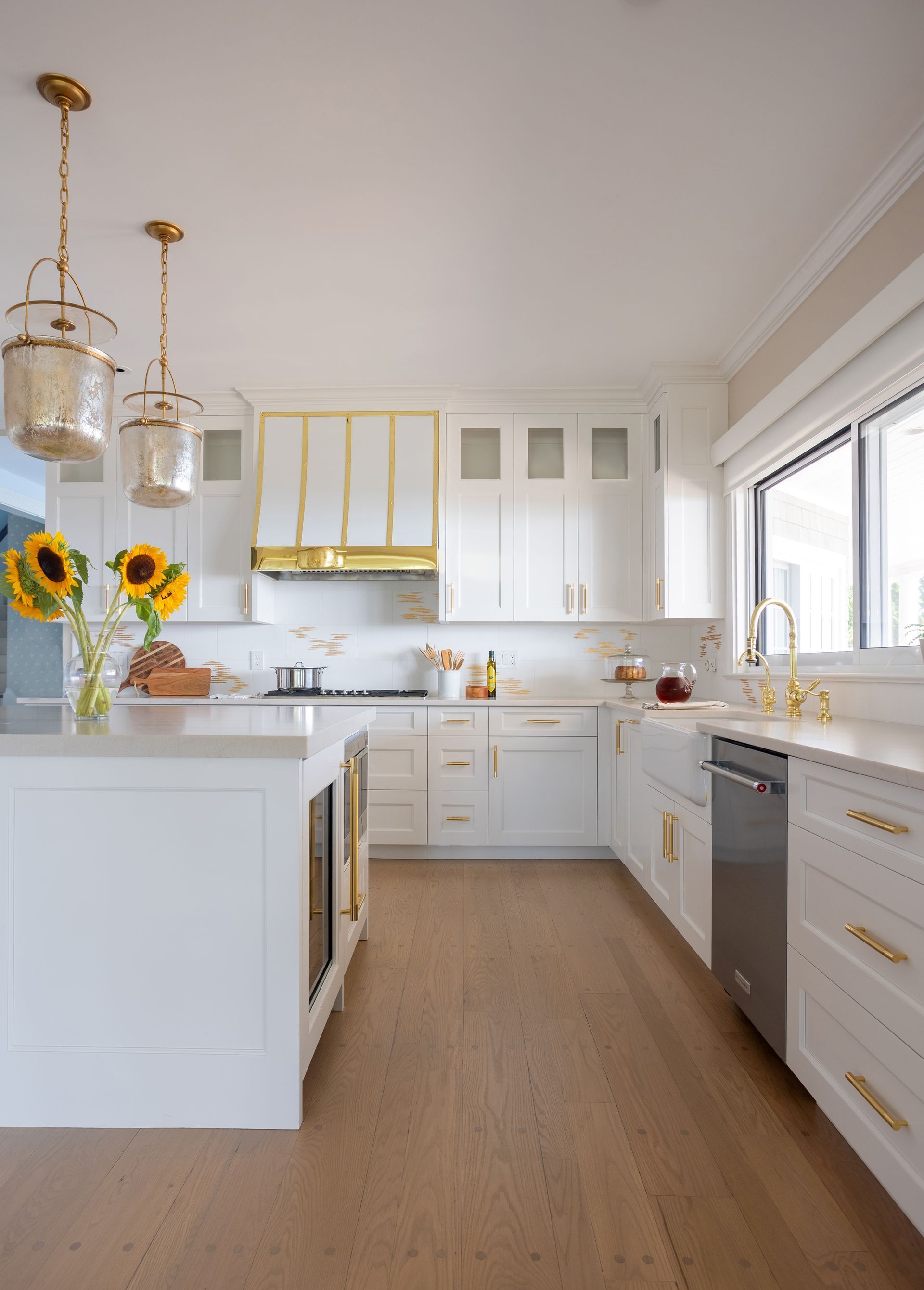Bright white kitchen with gold accents, sunflowers, and wood flooring, lit by natural light