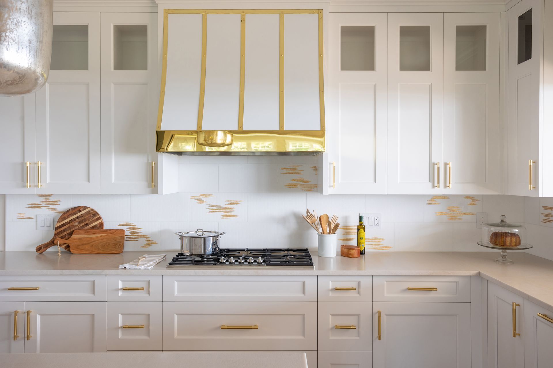 Bright white kitchen with gold accents, marble counters, and a gas stove centered under a gold range hood.