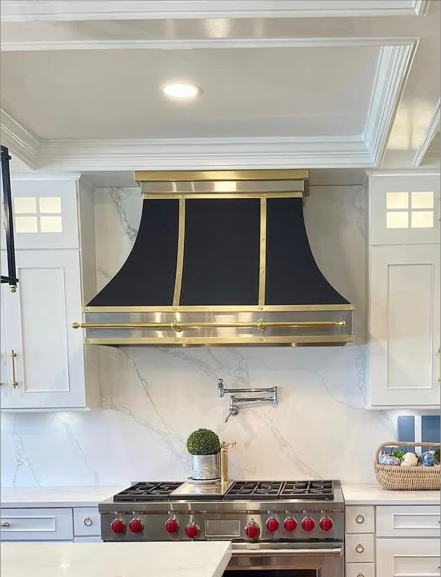 Elegant kitchen with black-and-gold range hood, stainless stove, white cabinets, and marble backsplash.