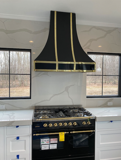 Black-and-gold range hood above a stove in a bright kitchen with windows and white cabinets