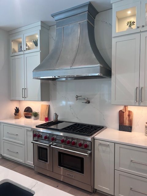 Modern white kitchen with stainless steel range and large hood, lit display cabinets, and countertop decor