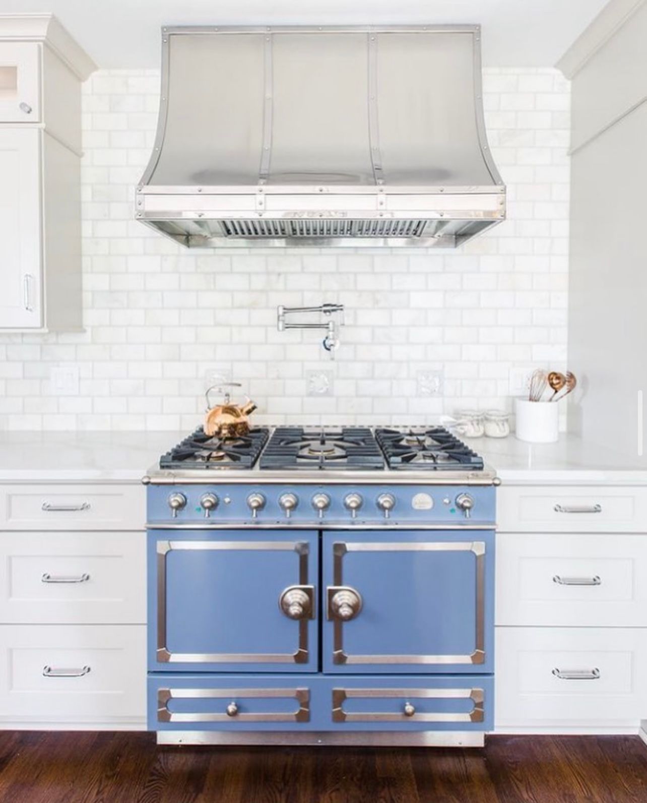 Bright blue vintage stove with stainless hood in a white tiled kitchen, flanked by white cabinets.