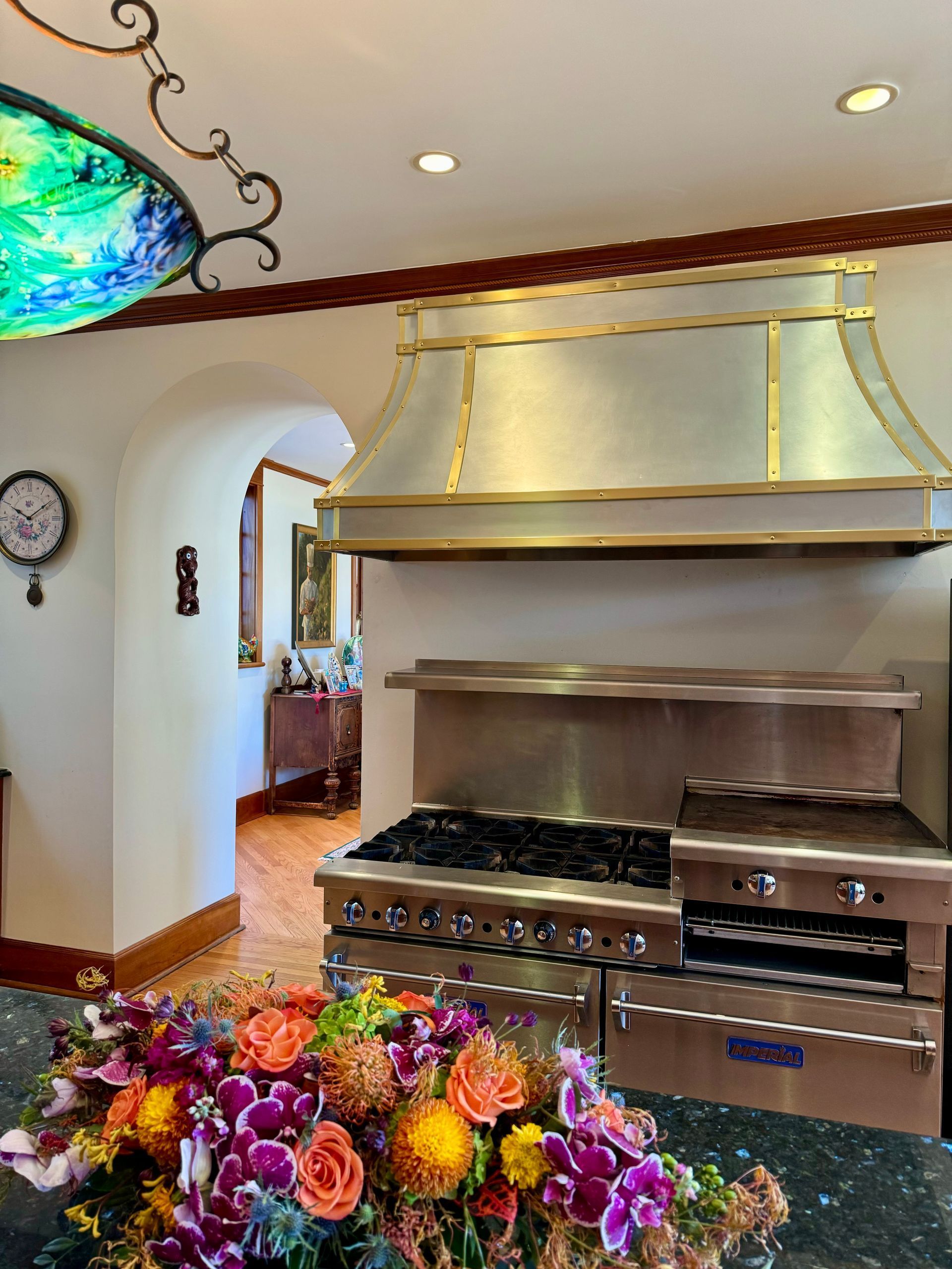 Elegant kitchen with stainless steel range, gold hood, arched doorway, and colorful flowers on a counter