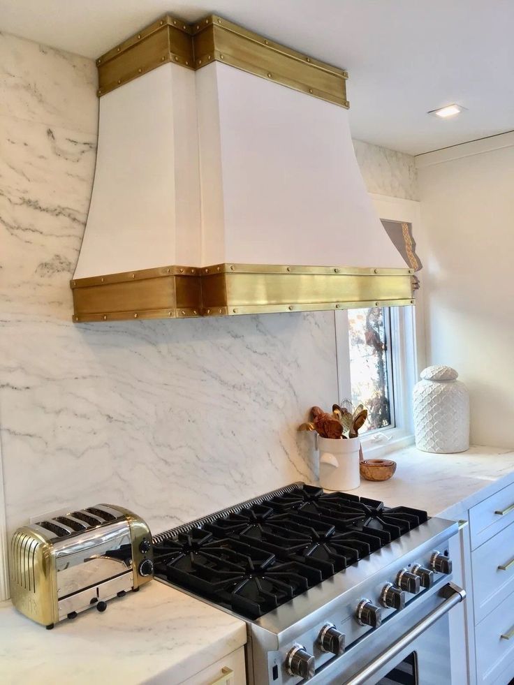 Modern kitchen with white marble walls, gold-trimmed vent hood, and stainless steel gas range