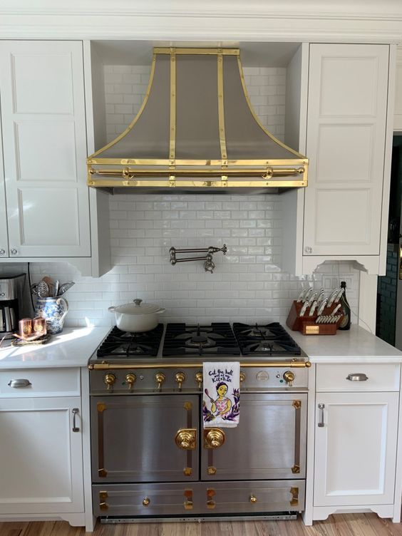Classic kitchen with white cabinets, stainless stove, and gold range hood against white subway tile.