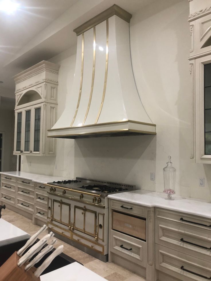 Elegant white and gold kitchen with a large range hood, marble counters, and built-in cabinetry