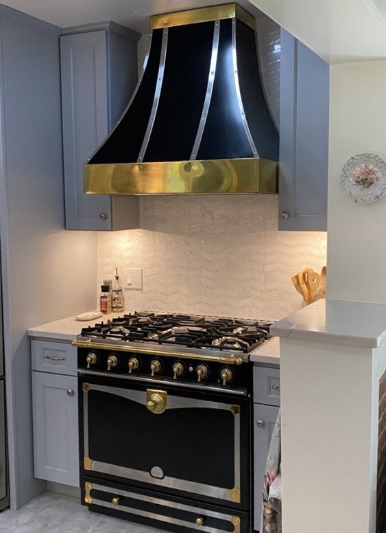 Elegant black-and-gold stove under a glossy black range hood in a blue-gray kitchen.