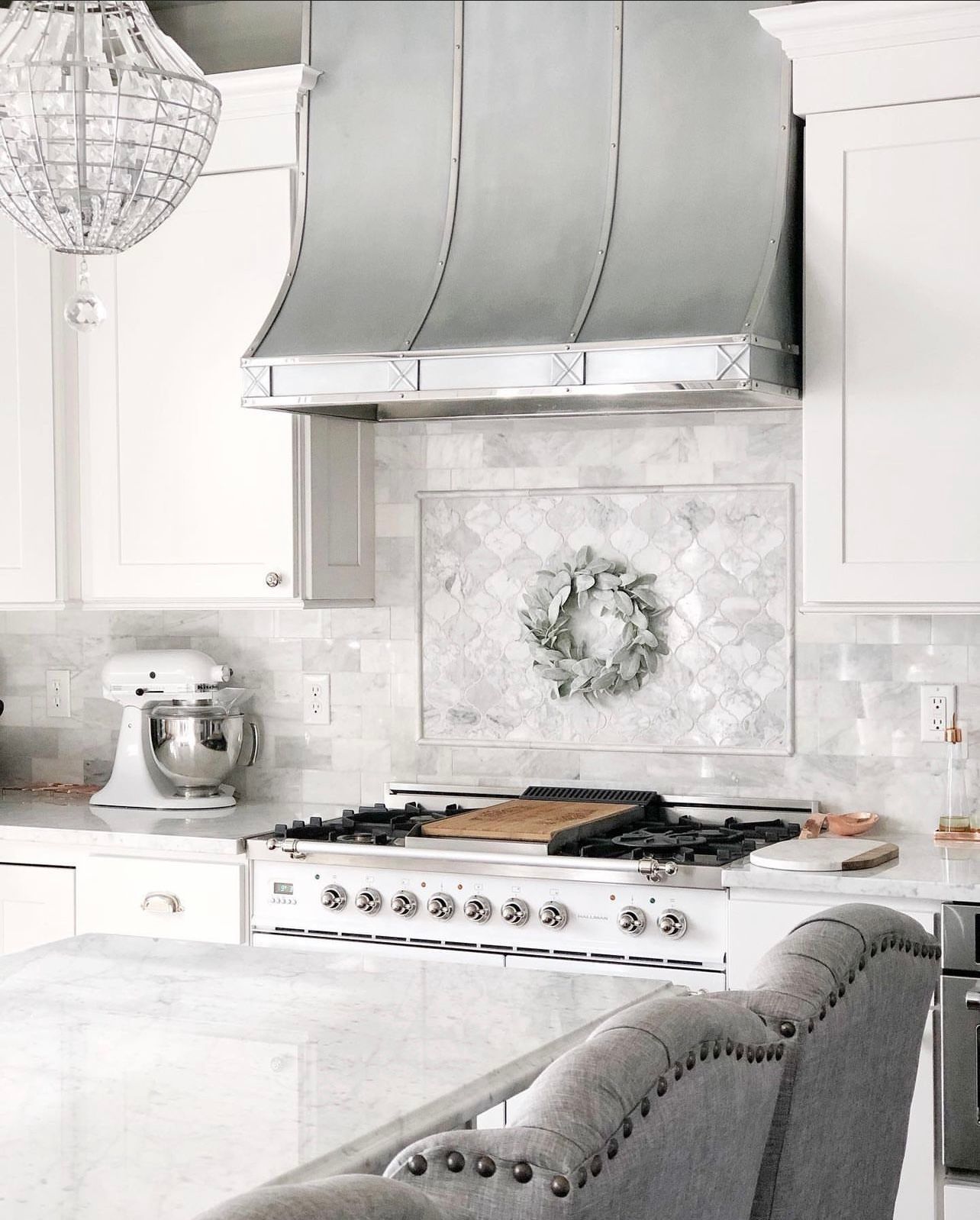 Bright white kitchen with gray range hood, marble backsplash, gas stove, and tufted gray stools.
