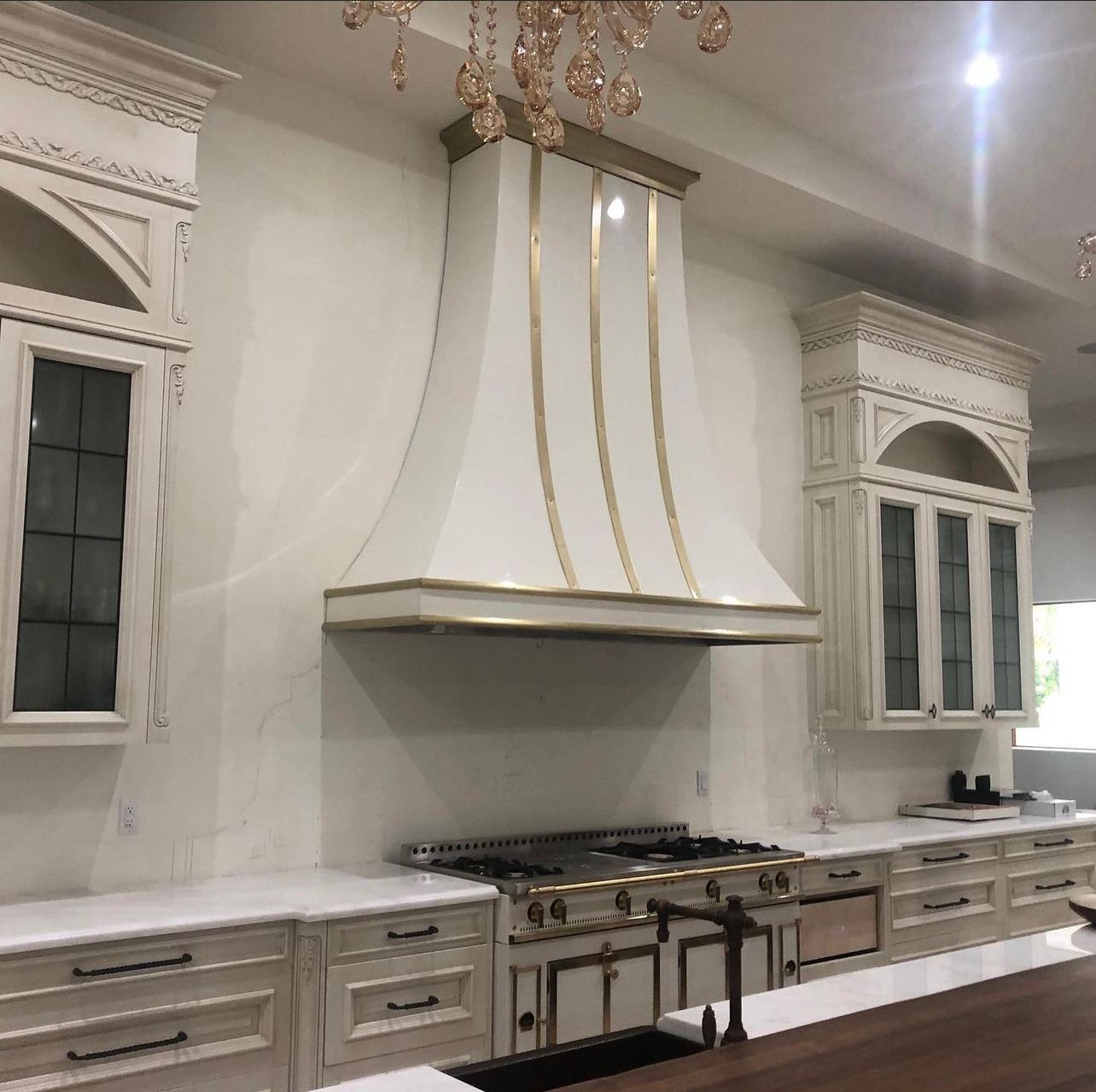 Elegant white kitchen with gold accents, large hood, marble counters, and ornate cabinets