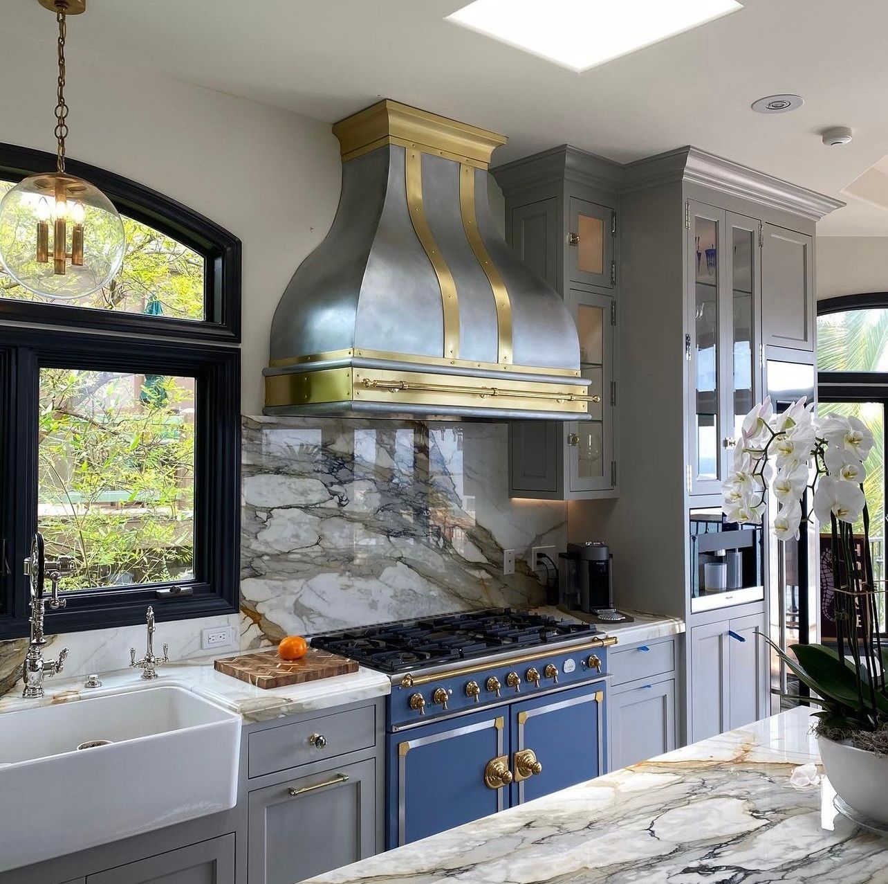 Elegant kitchen with blue range, gold hood, marble counters, black windows, and white cabinets