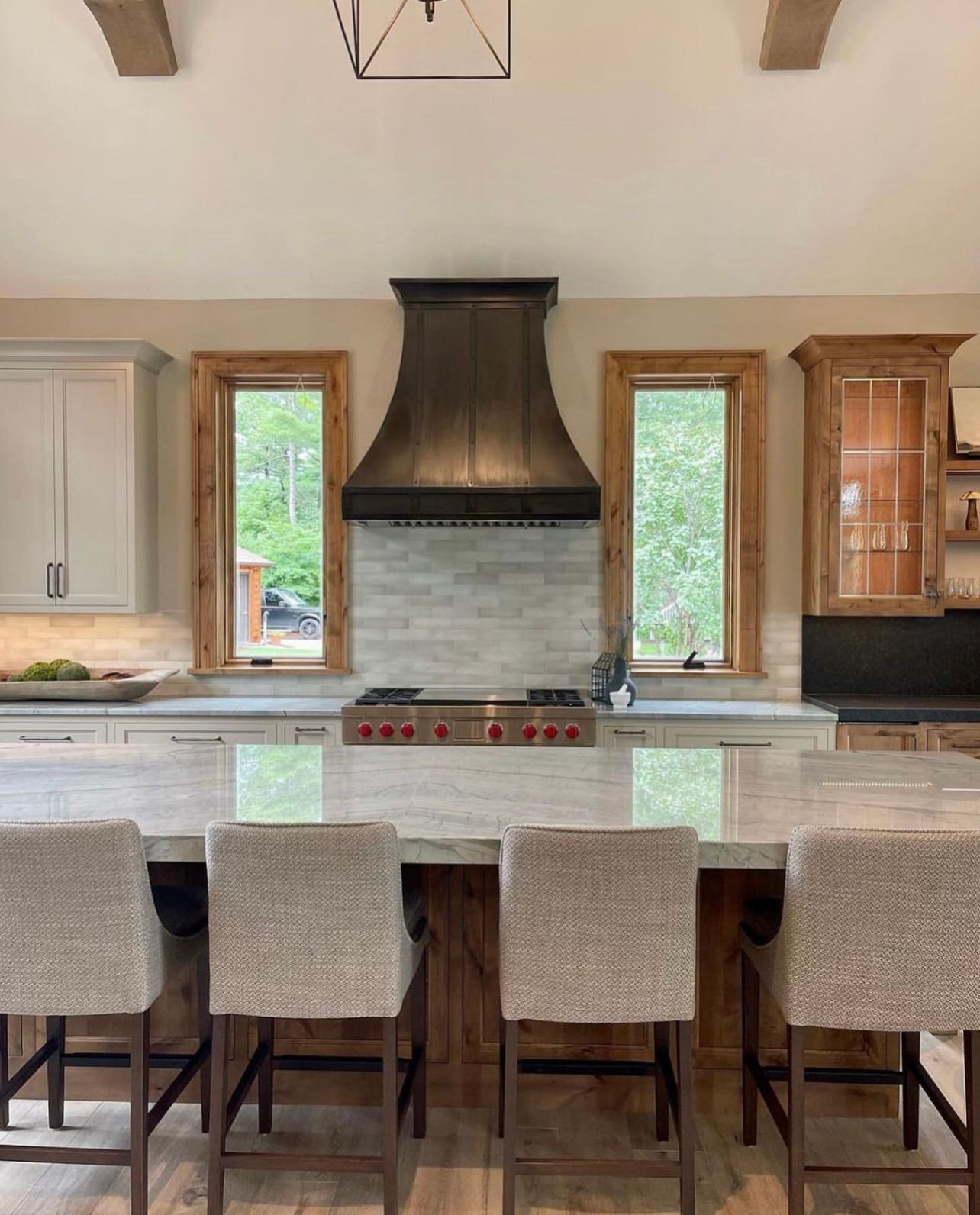 Modern kitchen with white island, six upholstered stools, and a black range hood centered between tall windows.