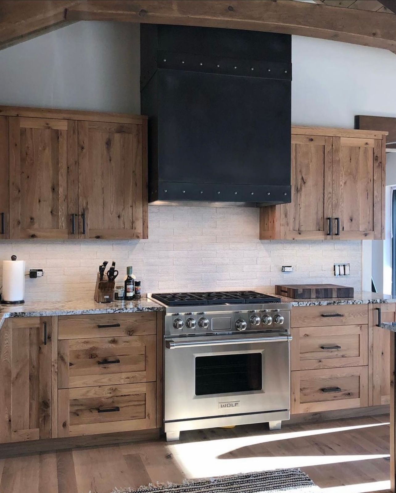 Rustic kitchen with wood cabinets, stainless steel range, and black vent hood beneath a vaulted ceiling.