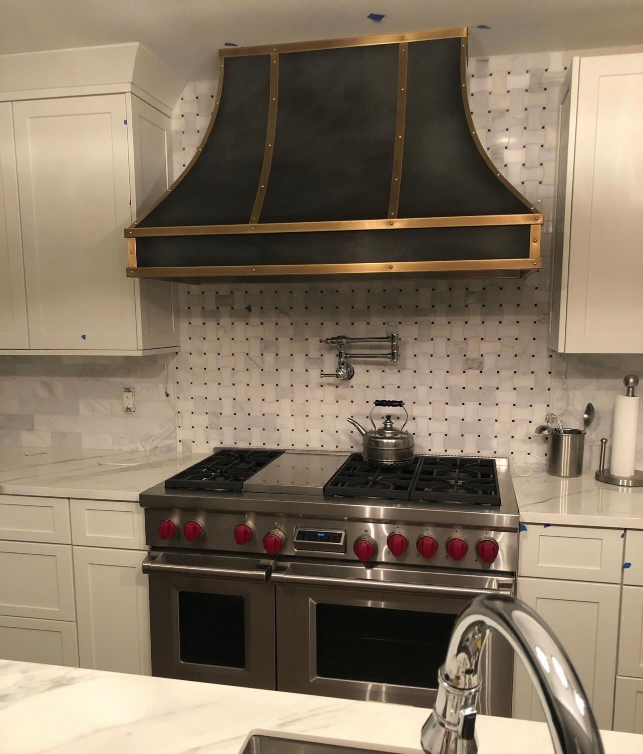White kitchen with a stainless steel range, red knobs, and a large black hood over a patterned tile backsplash.