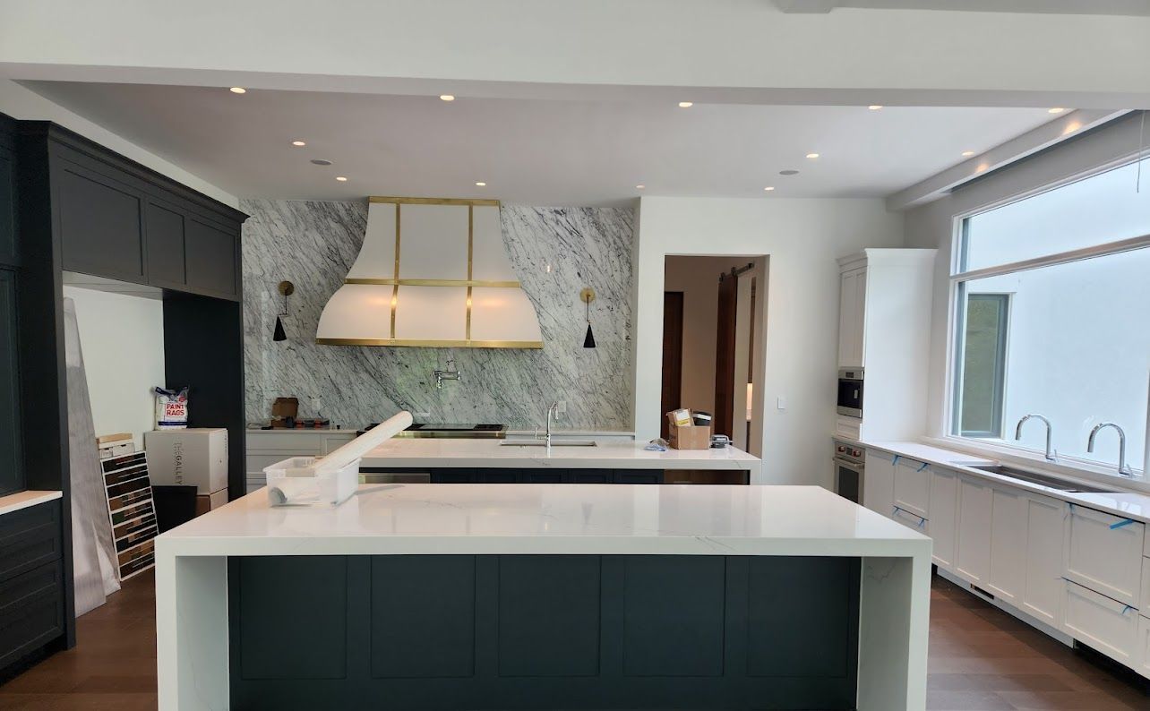 Modern kitchen with a large island, white countertops, dark cabinetry, and a marble backsplash by a bright window.