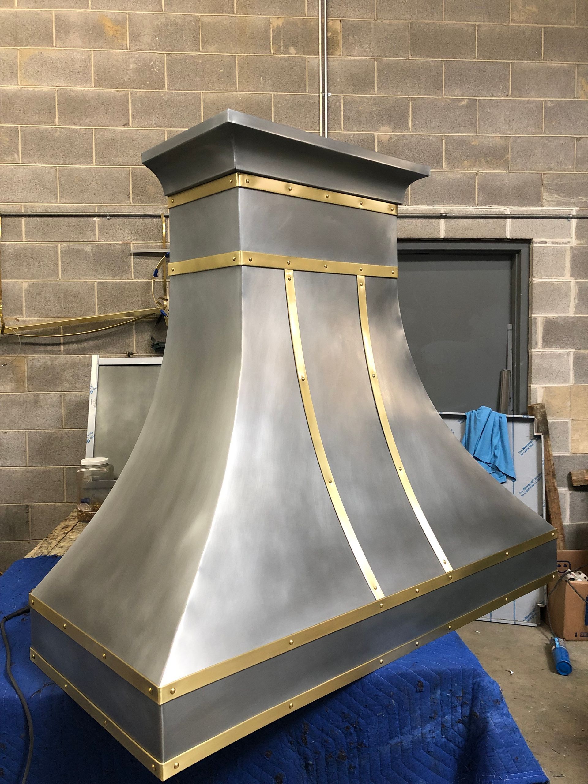 Metal chimney hood with brushed steel finish, gold trim, against a brick wall in a workshop