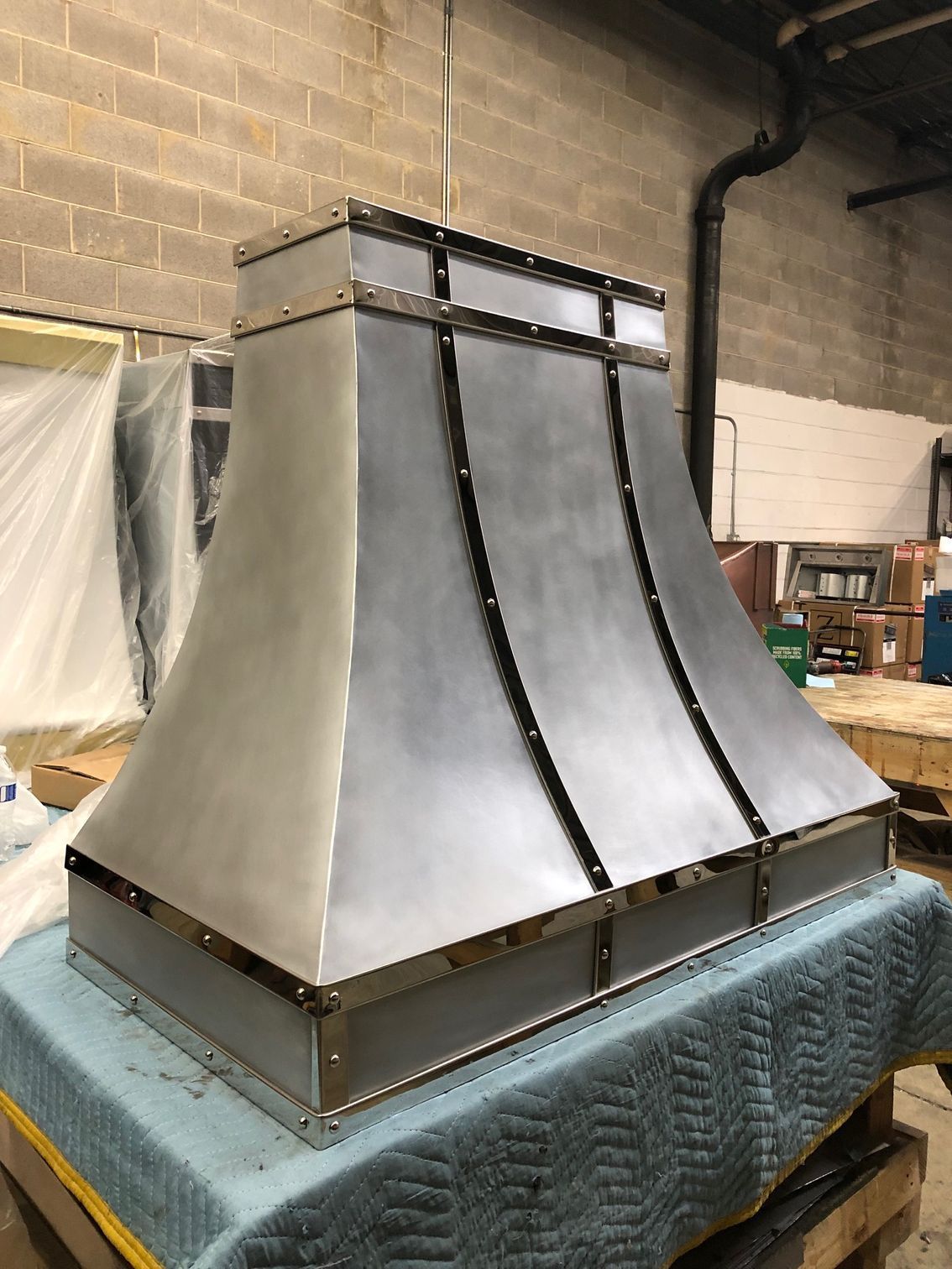 Metal industrial hood on a padded worktable in a workshop, with brick walls and wrapped equipment in the background