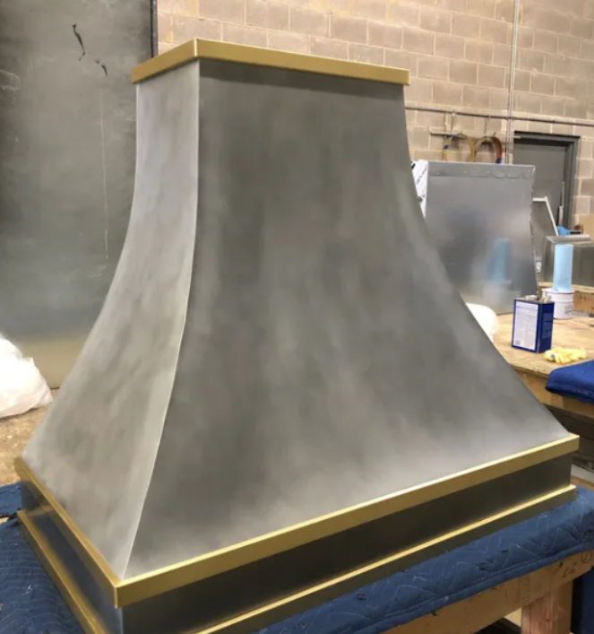 Large gray tapered metal hood with gold trim, displayed in a workshop on a blue-covered table