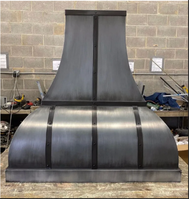 Large black metal industrial hood or duct assembly standing in a workshop against a cinderblock wall