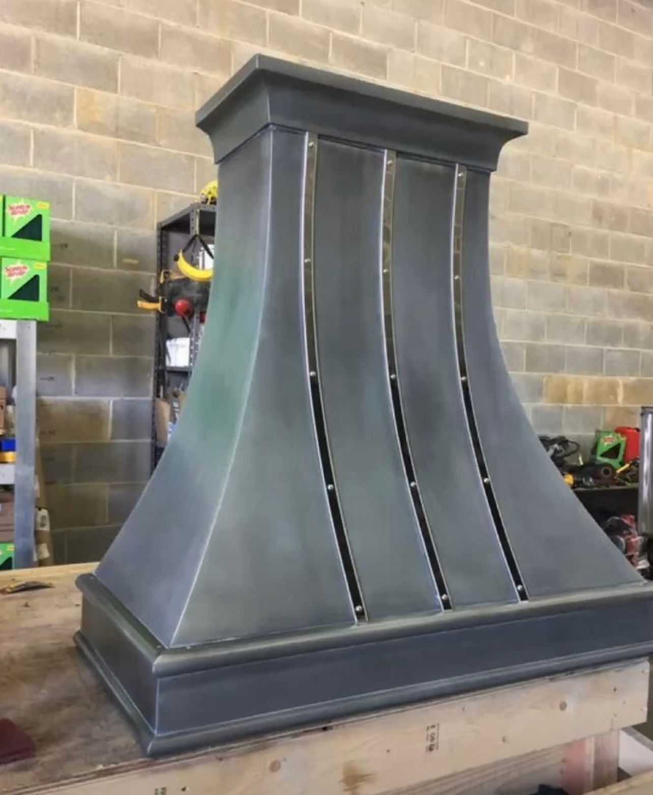 Large gray metal chimney hood with flared sides, standing in a workshop against a cinderblock wall