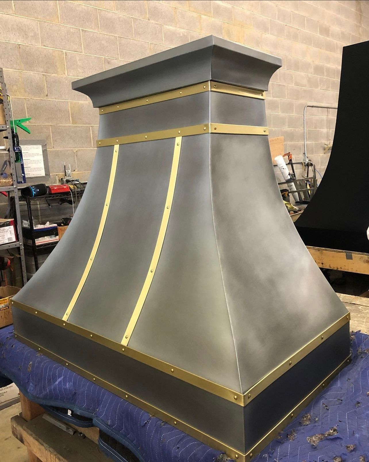 Gray metal chimney hood with gold trim, sitting in a workshop on a blue-covered pallet
