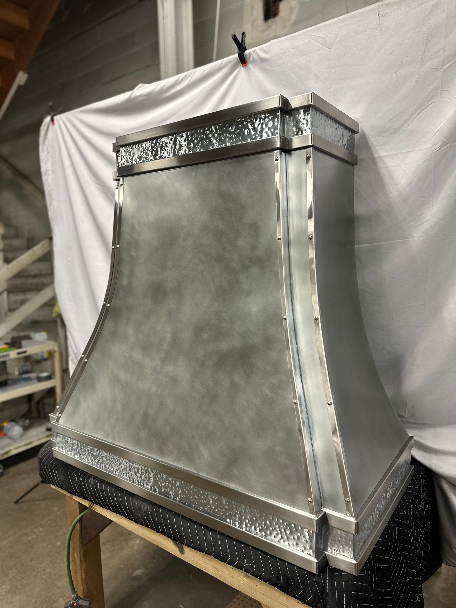 Metallic silver vent hood or chimney component on a workbench in a workshop, with decorative trim.