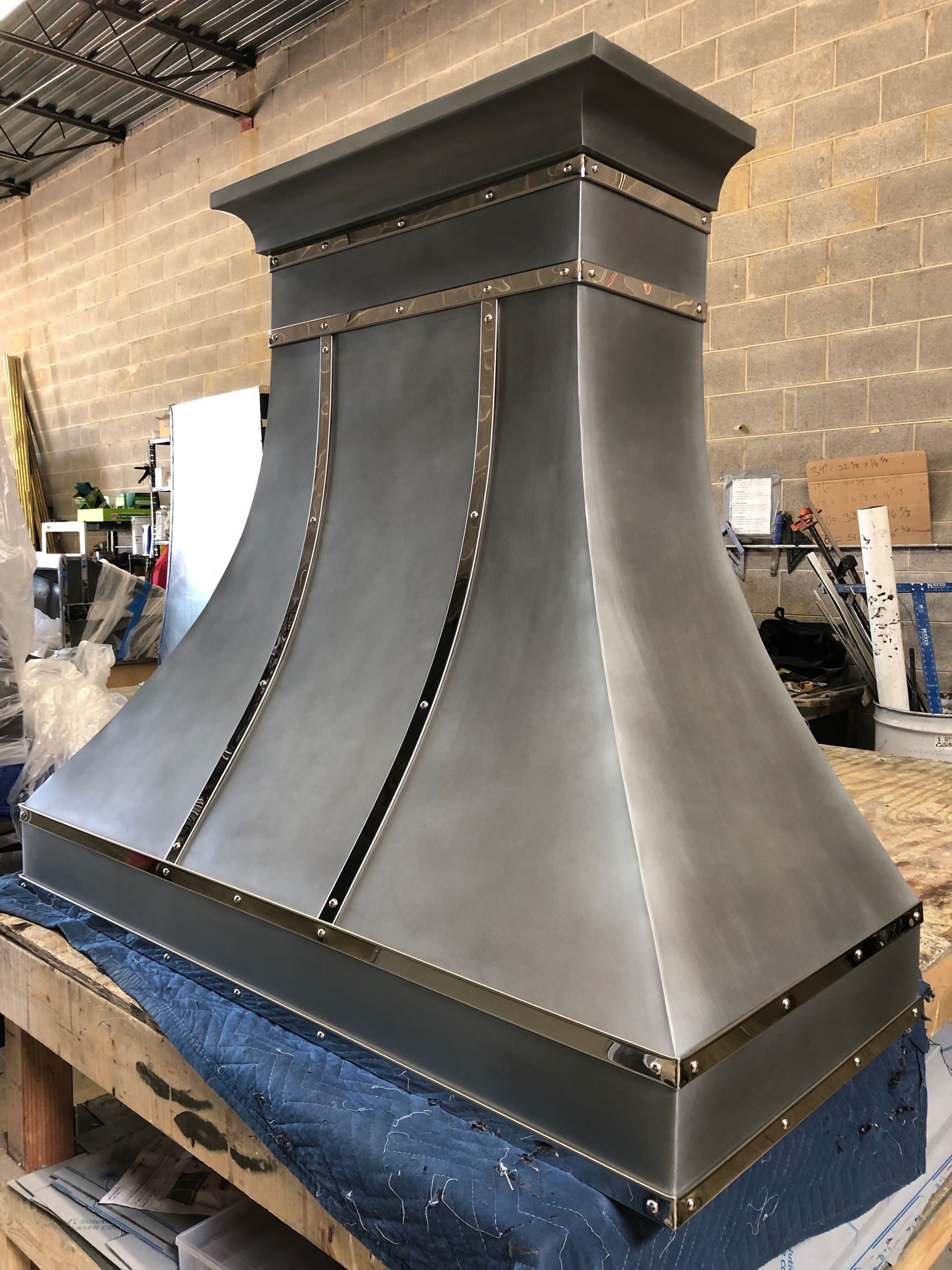 Large dark metal range hood with riveted seams, sitting in a workshop on a blue-covered base.
