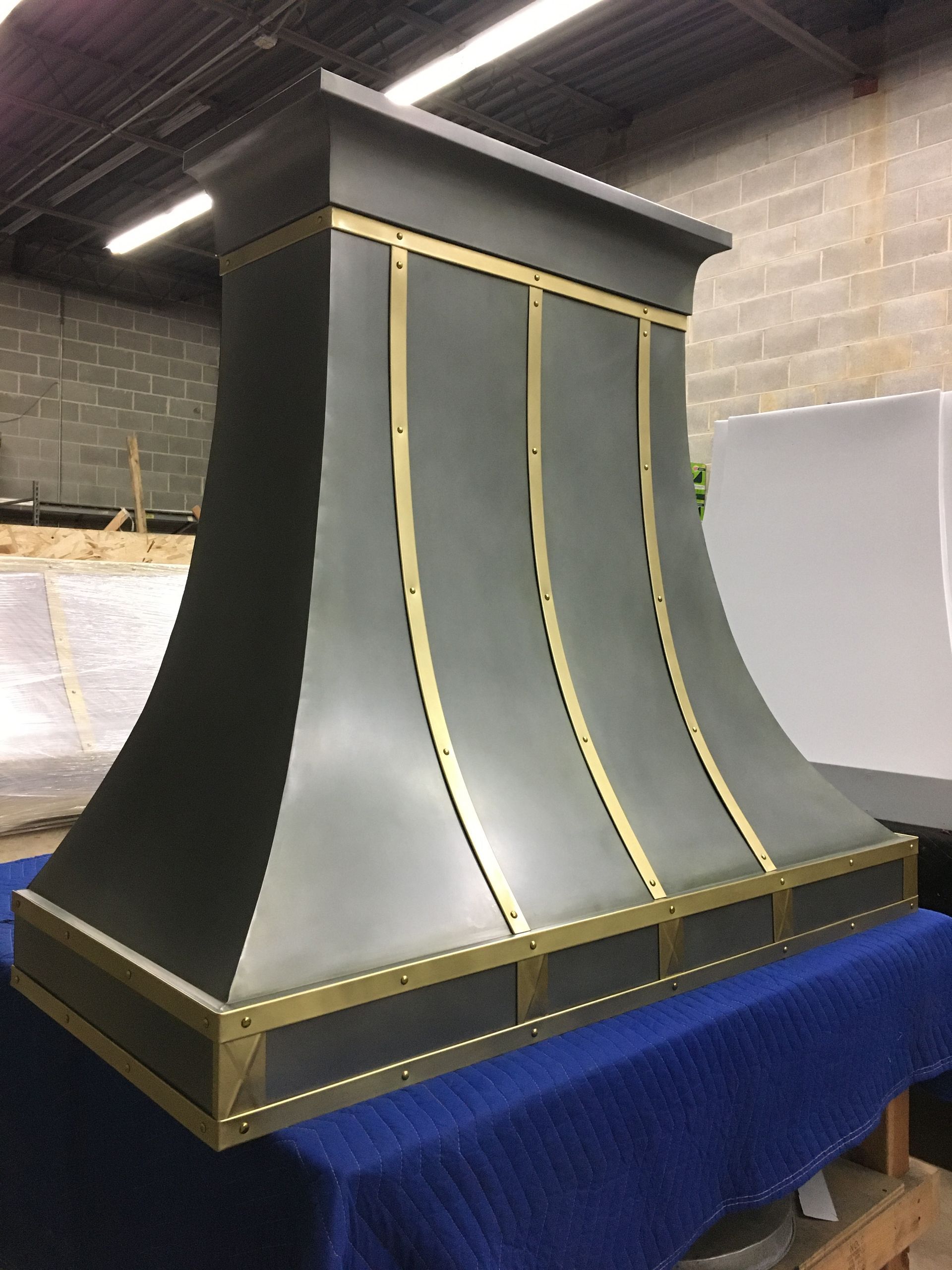 Decorative black and gray metal hood or vent with gold trim on a blue cloth-covered table in a workshop