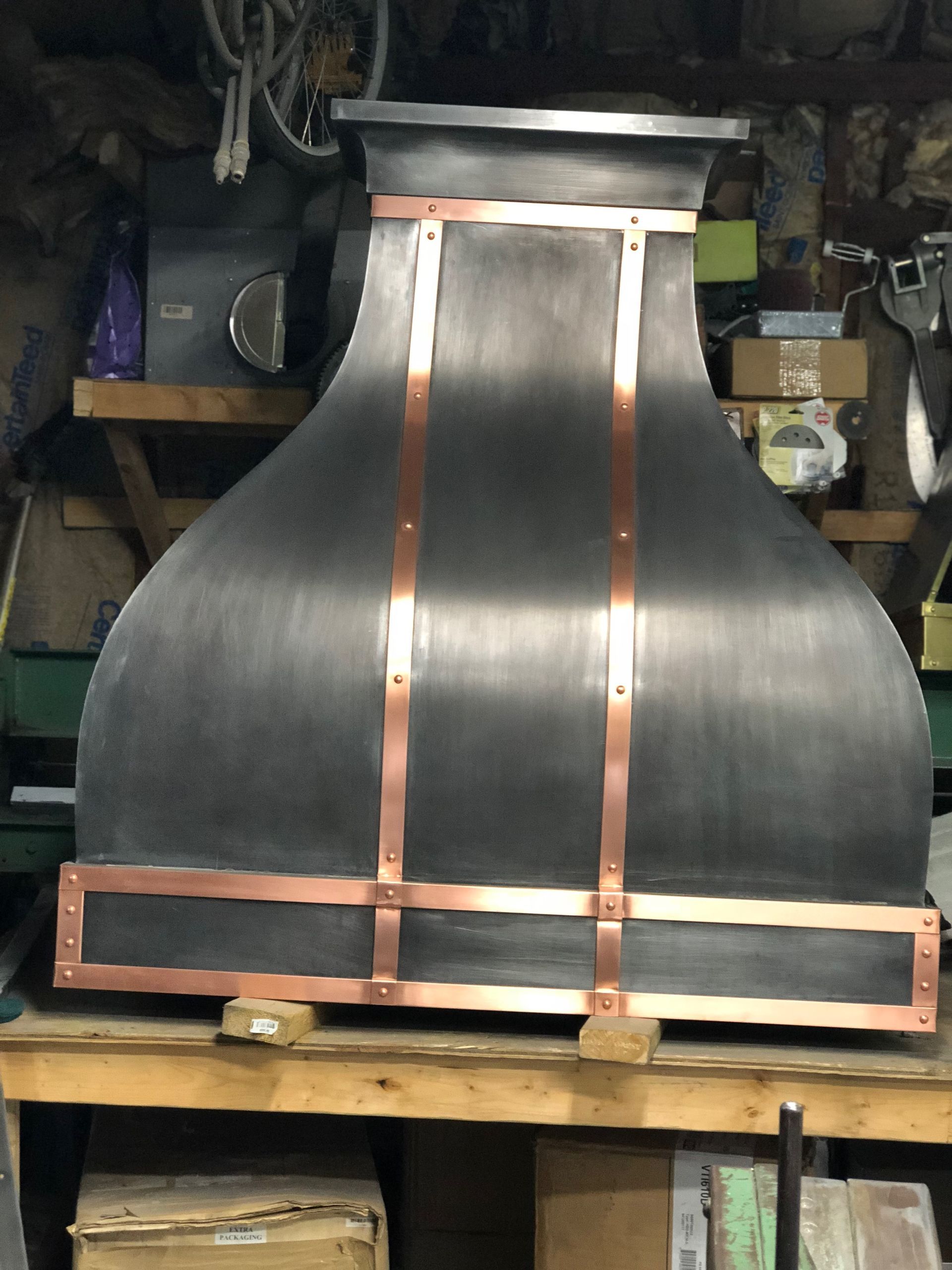 Large black metal hood with copper supports on a workbench in a cluttered workshop