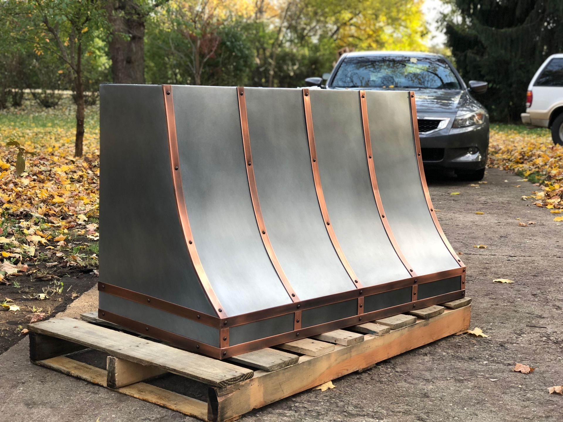 Curved metal sculpture on a wooden pallet outdoors, with a car and autumn trees in the background