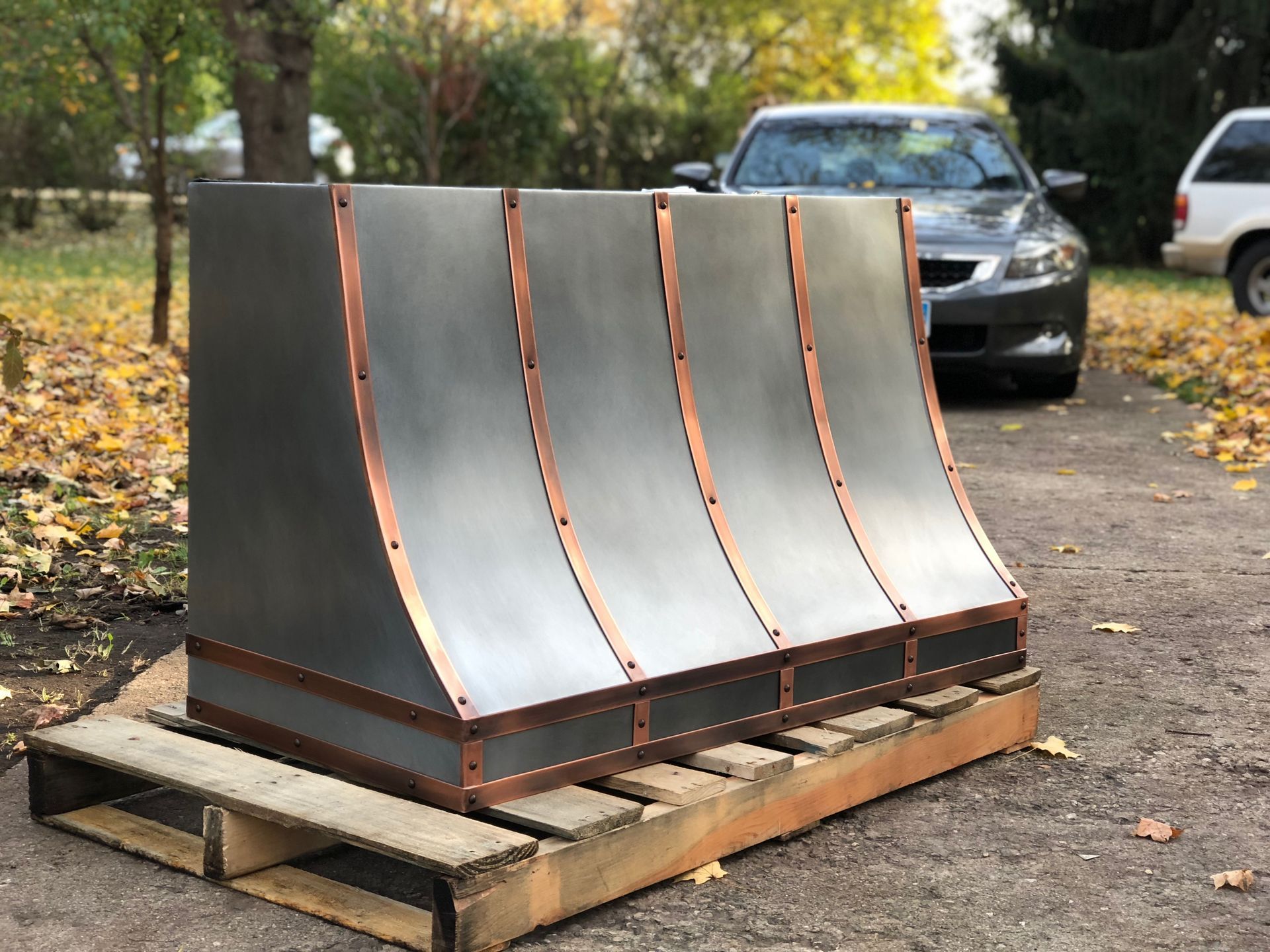 Four curved metal panels mounted on a wooden pallet outdoors, with a car in the background.