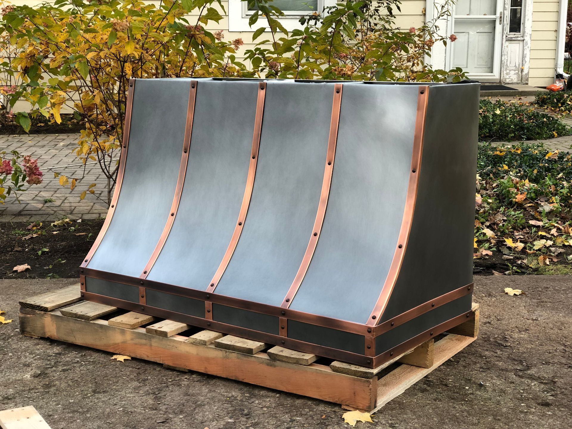 Outdoor metal panel sculpture on a wooden pallet, with curved gray panels and copper-colored edges.