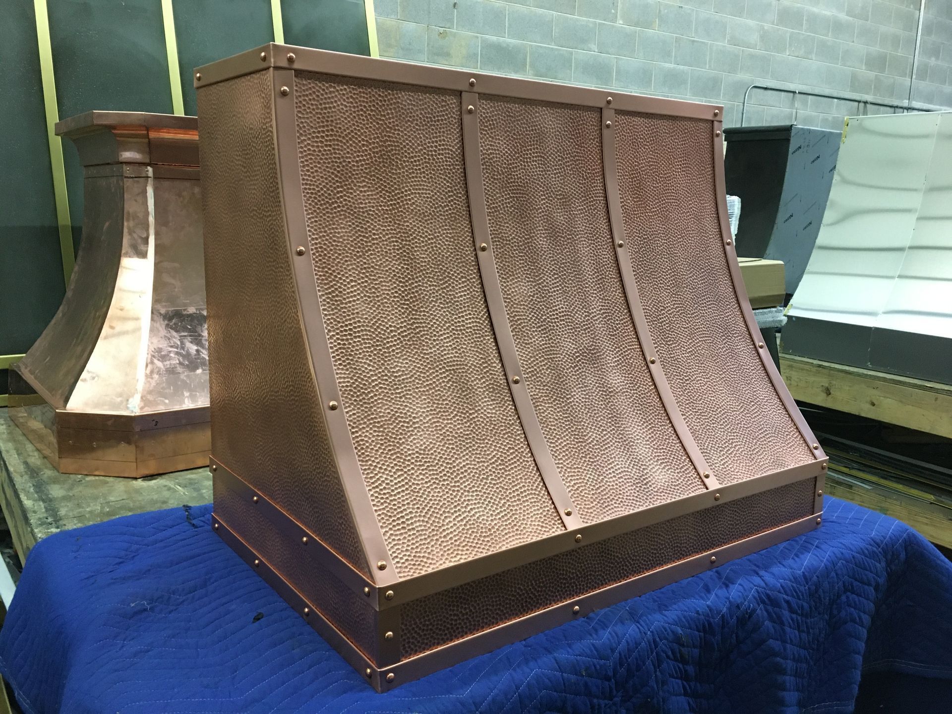 Copper range hood with three angled panels on a blue cloth in a workshop-like room