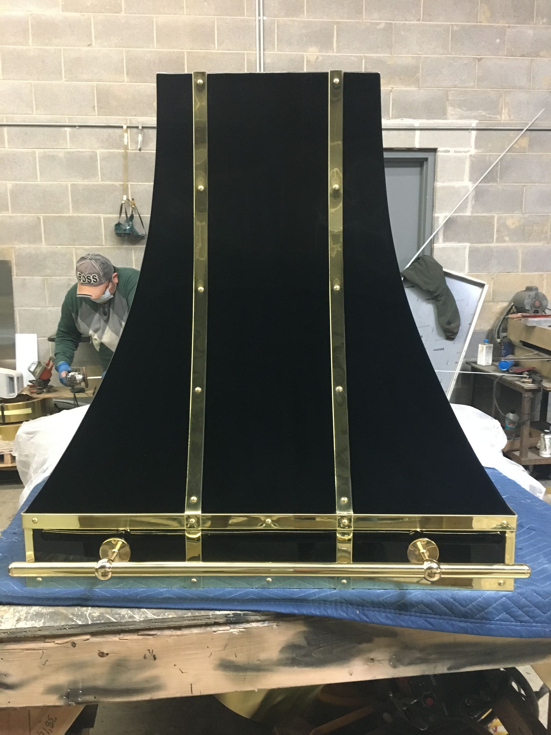 Large black industrial hood or chimney with brass trim in a workshop against a brick wall