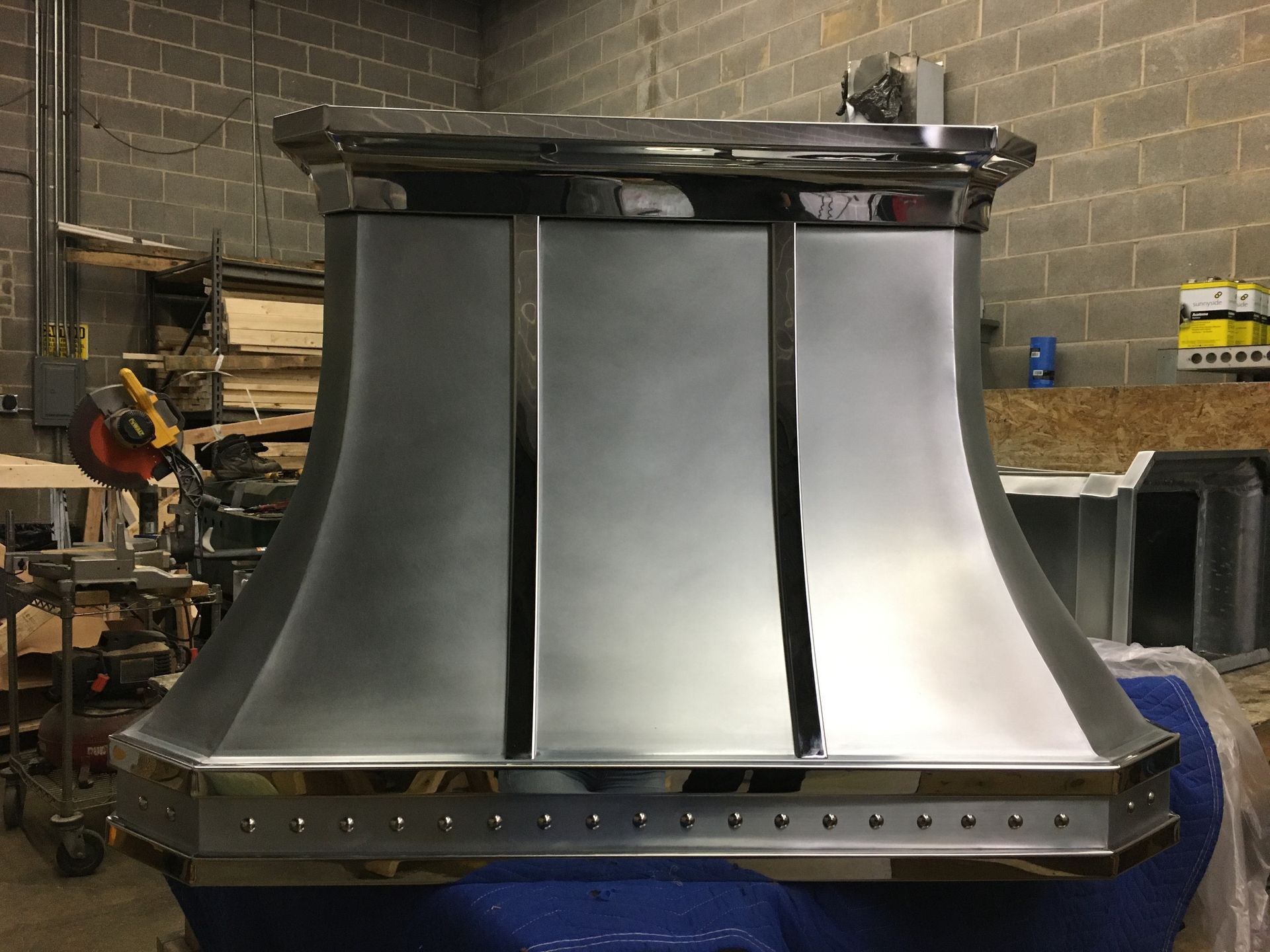 Large polished metal industrial hood in a workshop, with a row of rivets along the bottom.