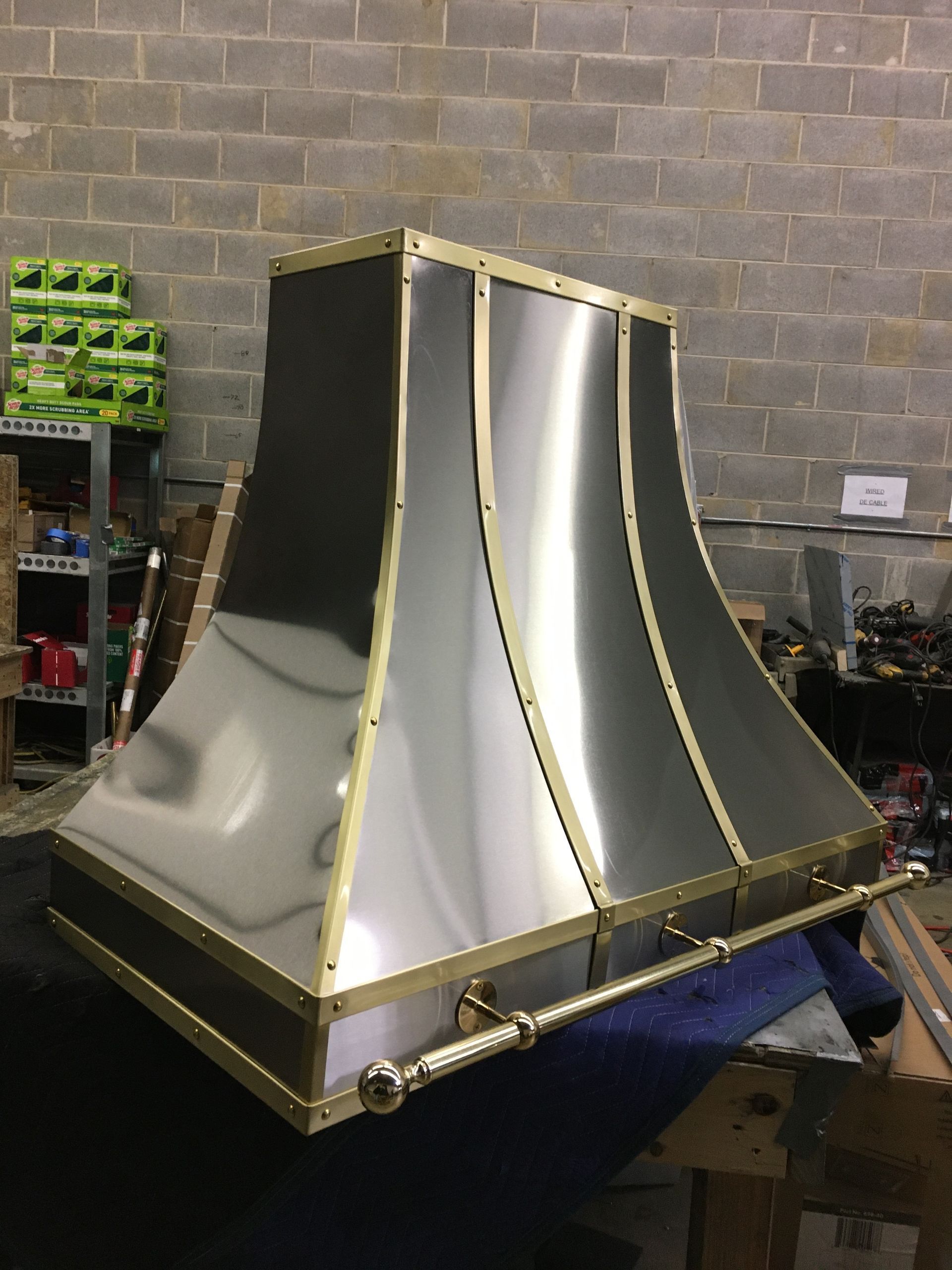 Metallic custom vent hood prototype on a workbench in a workshop against a brick wall.