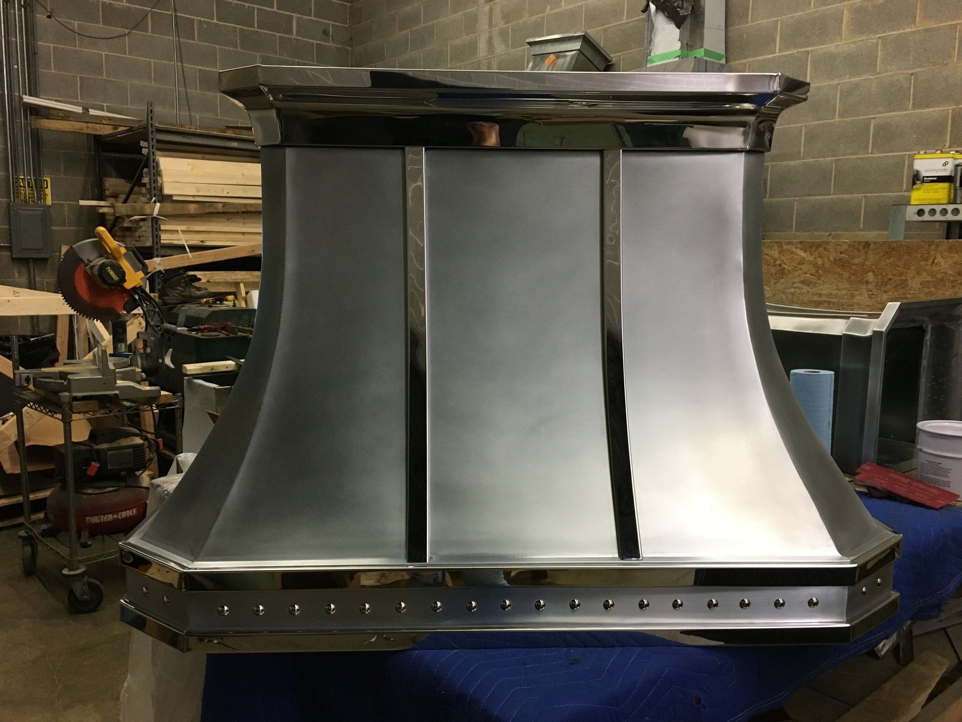 Large silver metal chimney hood in a workshop, with a black top and riveted base.
