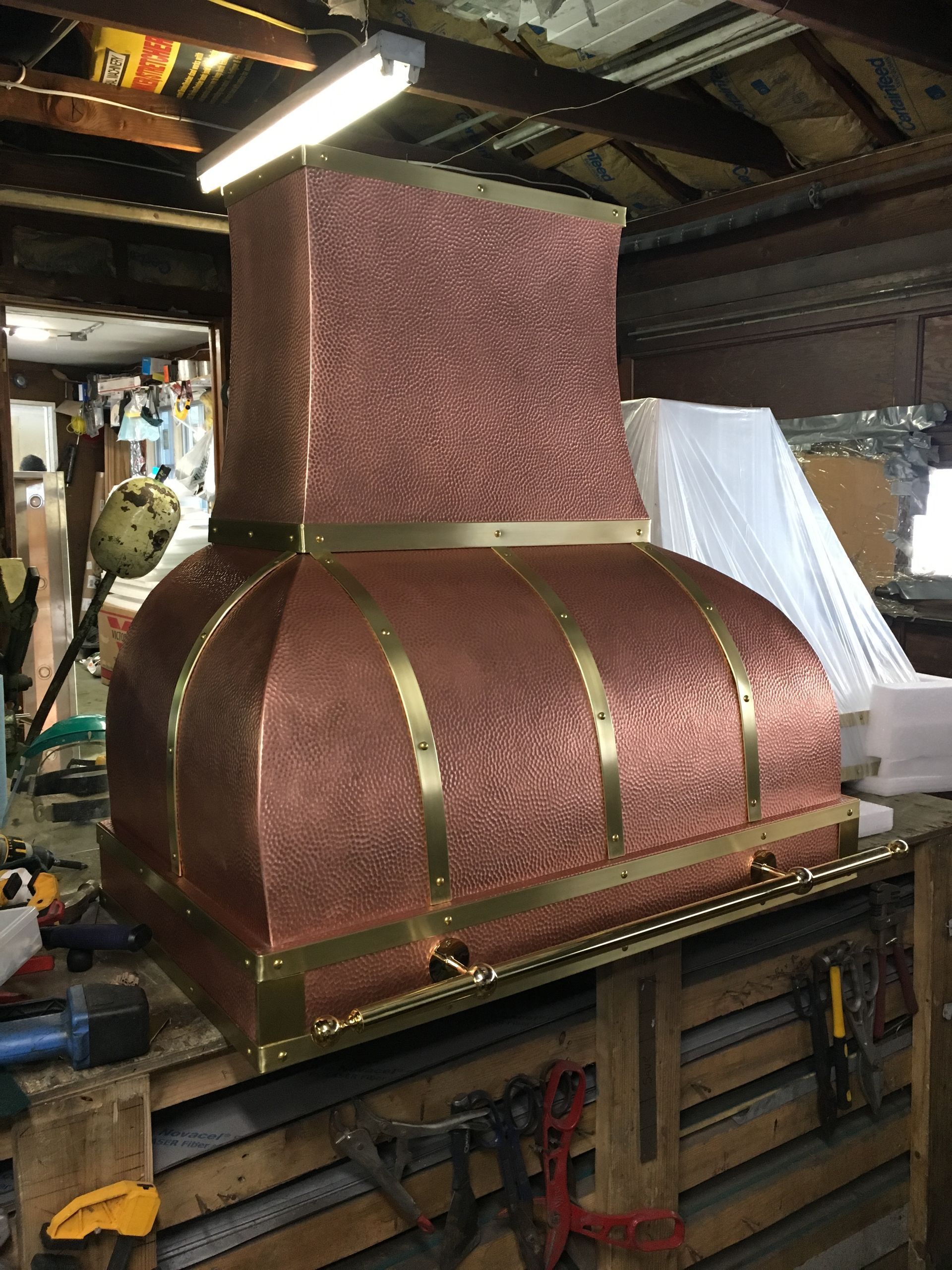 Copper-colored metal chimney hood in a workshop, with brass trim and a square vent stack.