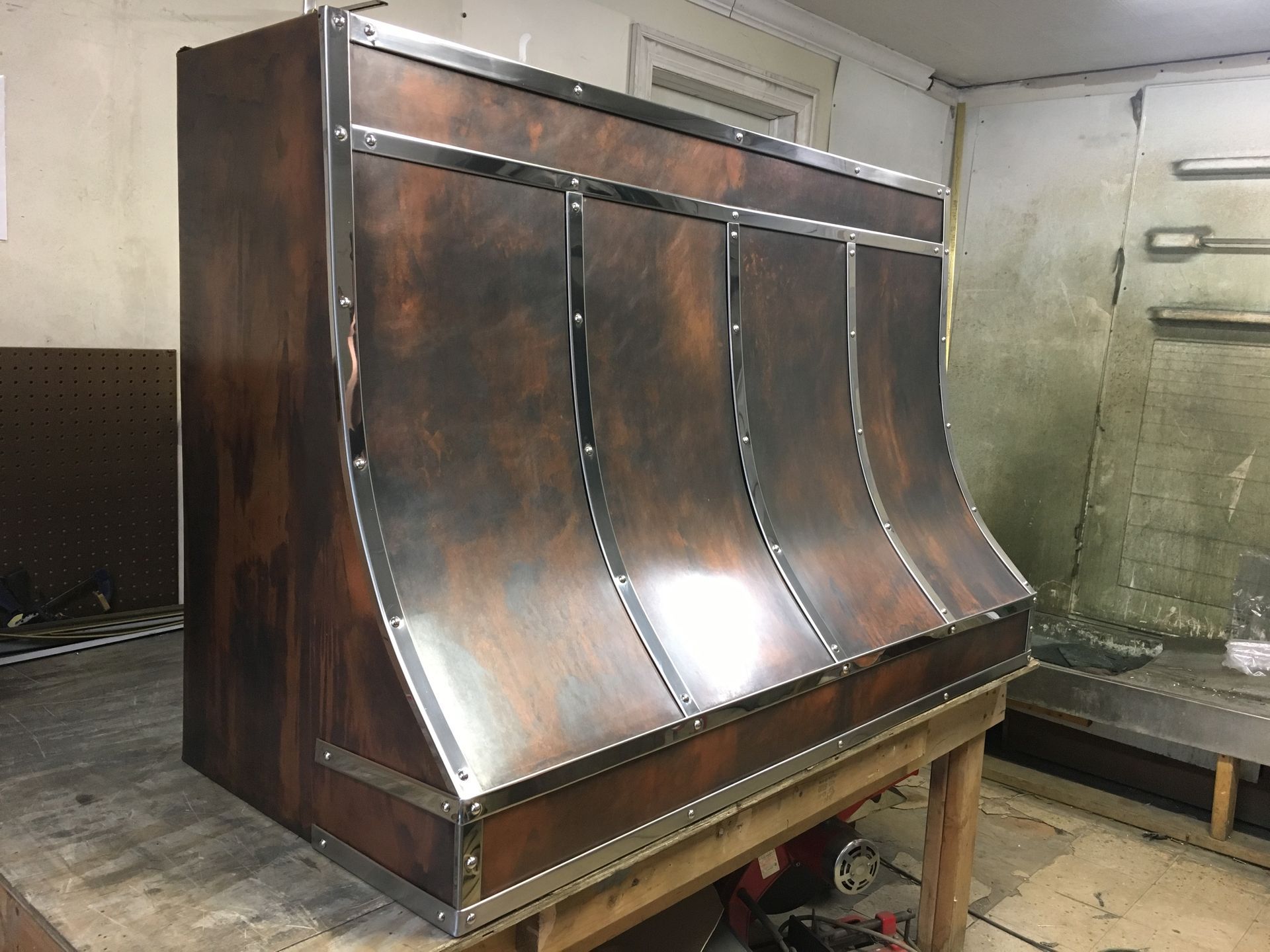 Large stainless steel industrial hopper or chute in a workshop, mounted on a wooden stand.