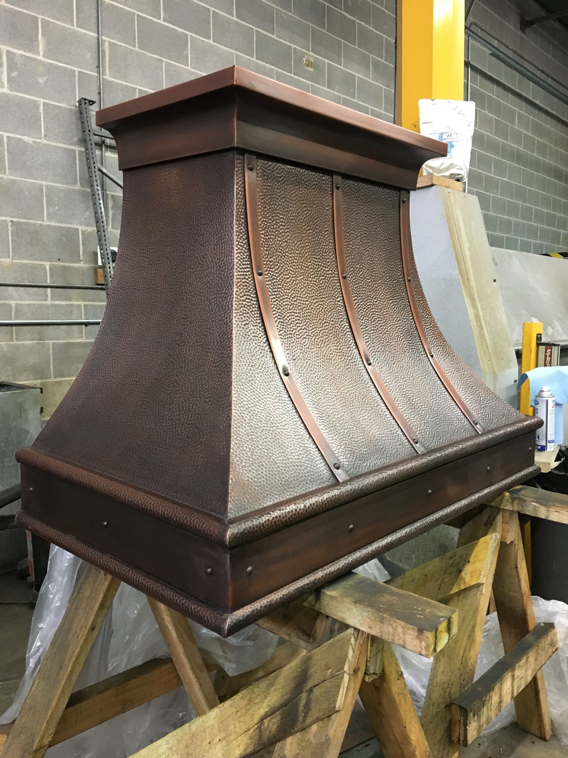 Copper chimney hood on a wooden stand in a workshop, with textured brown panels and a dark trim.