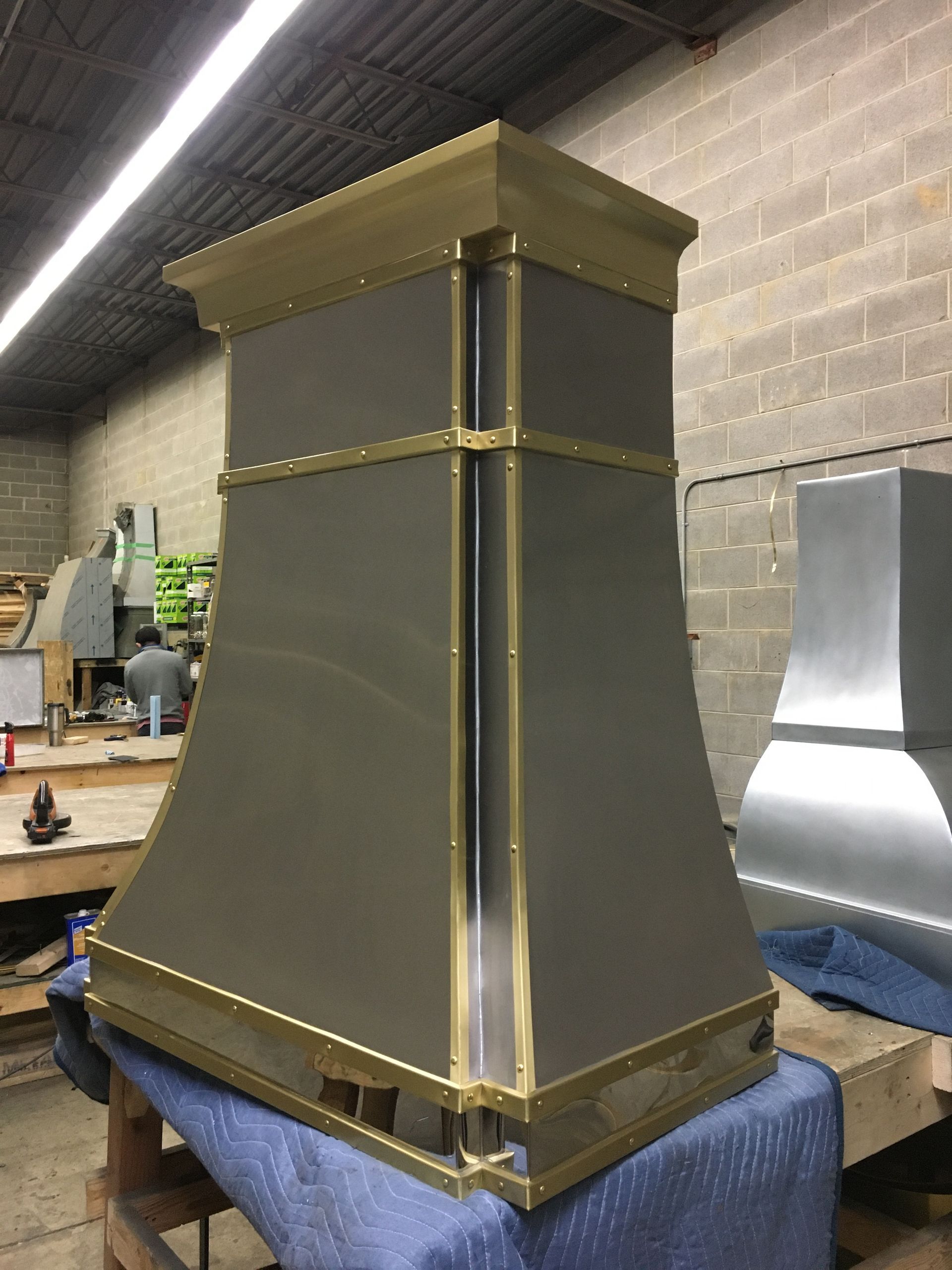 Brass-and-gray metal range hood on a workbench in a workshop with tools and a brick wall backdrop