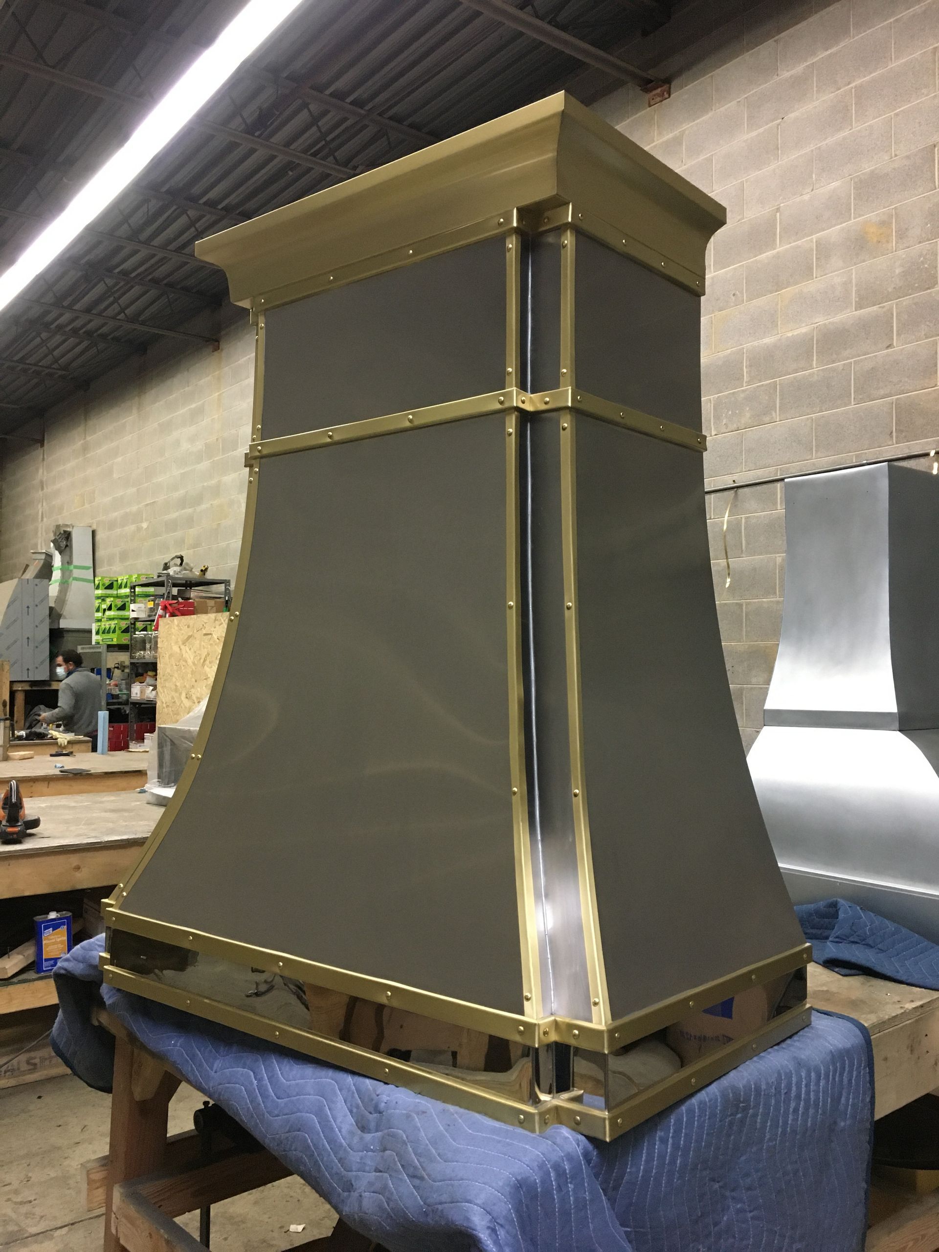Large gray metal chimney hood with gold trim, sitting on a blue blanket in a workshop