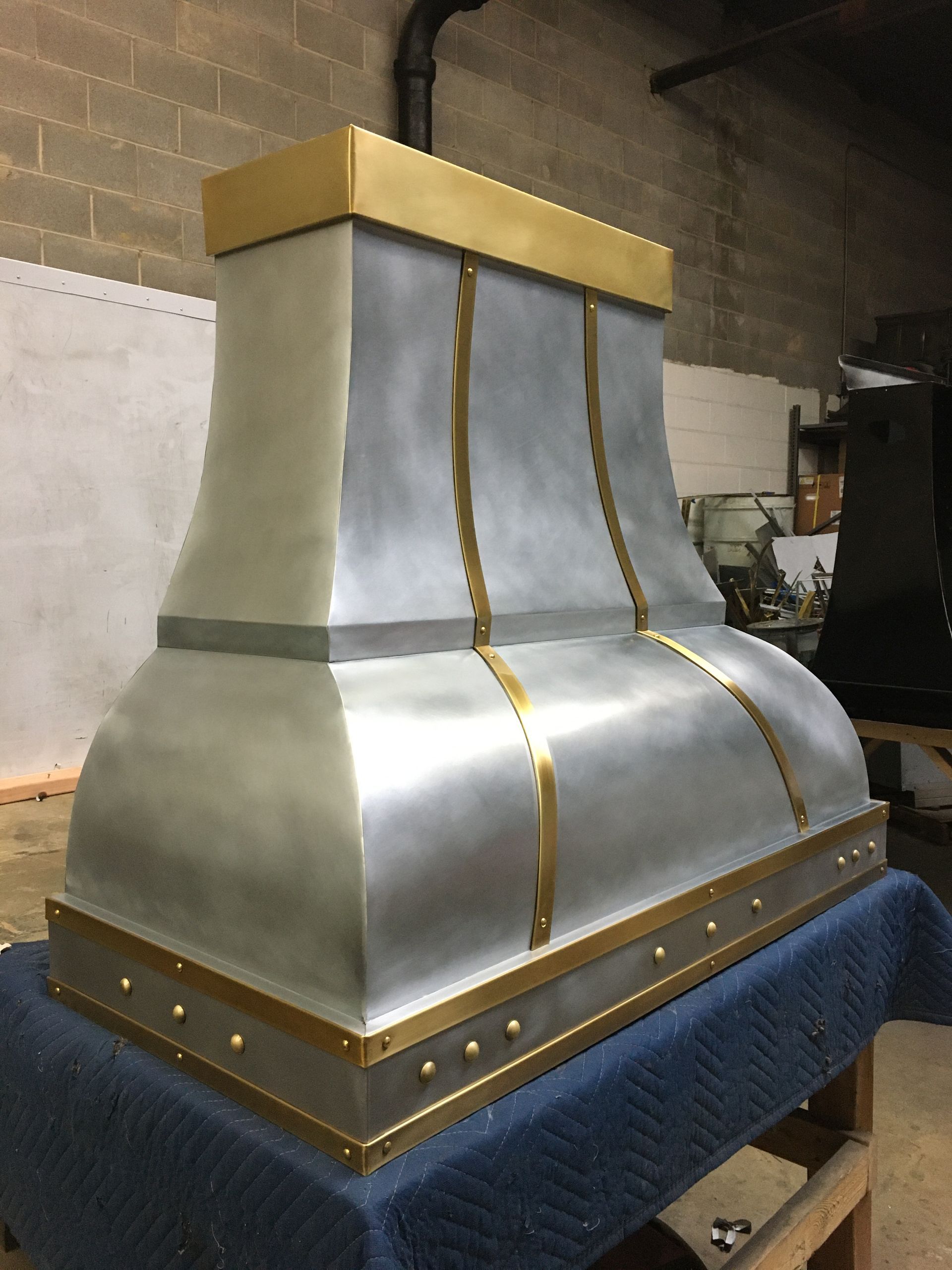 Silver metal ventilation hood with gold trim on a blue-covered table in a workshop.