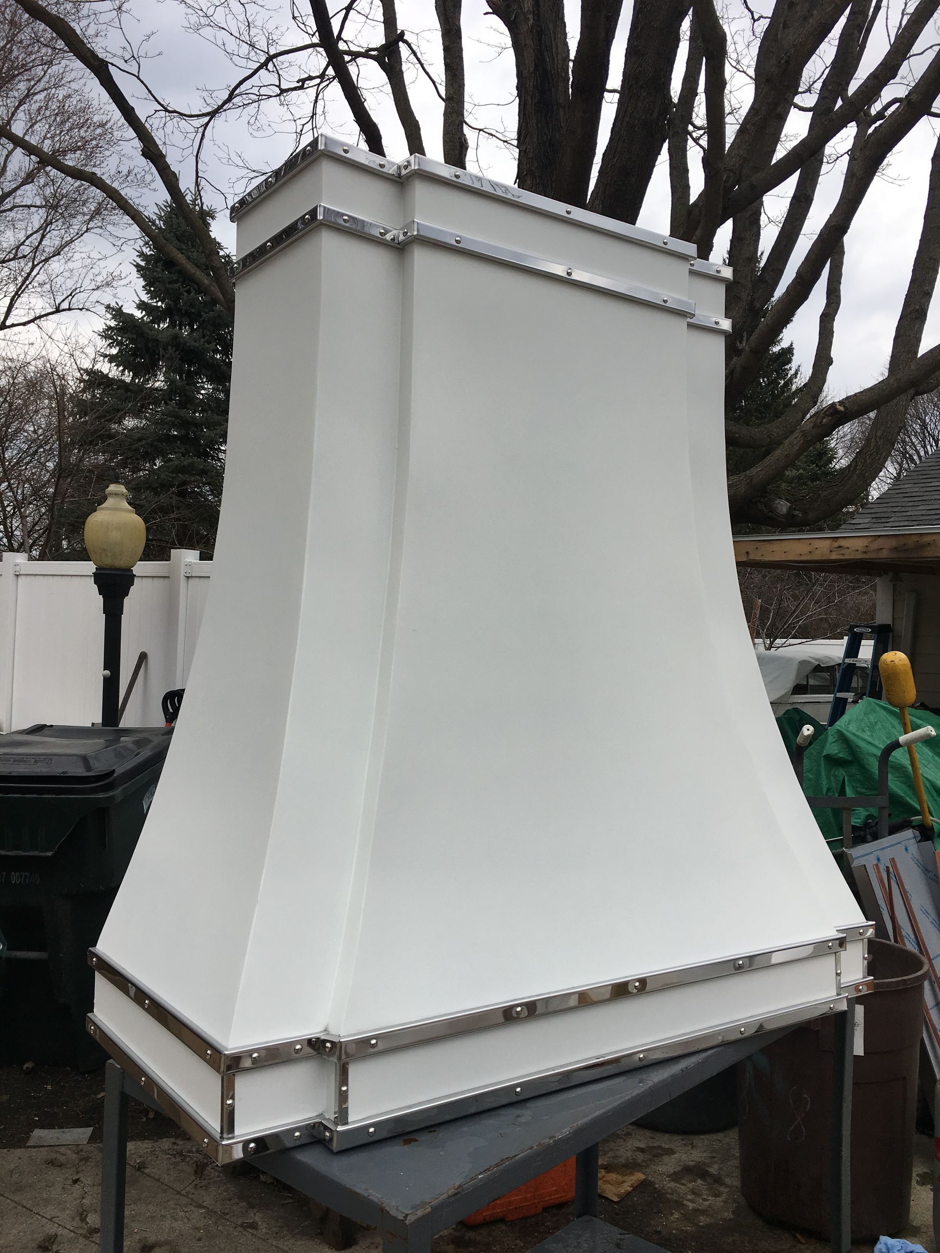 White decorative chimney hood on a metal base outdoors among trees and garden equipment