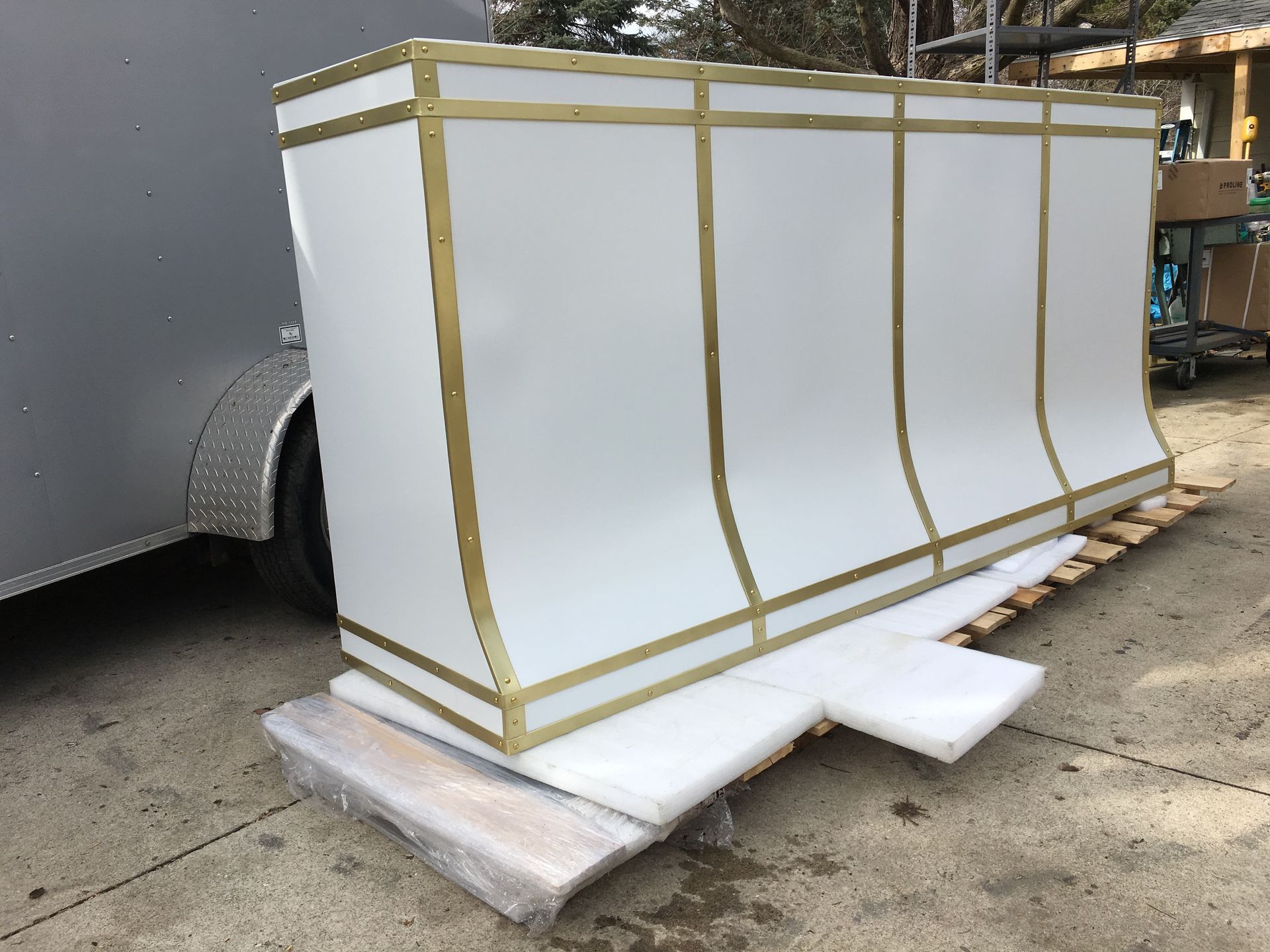 Large white crate or panel wrapped in tape, sitting on a pallet outdoors beside a trailer.