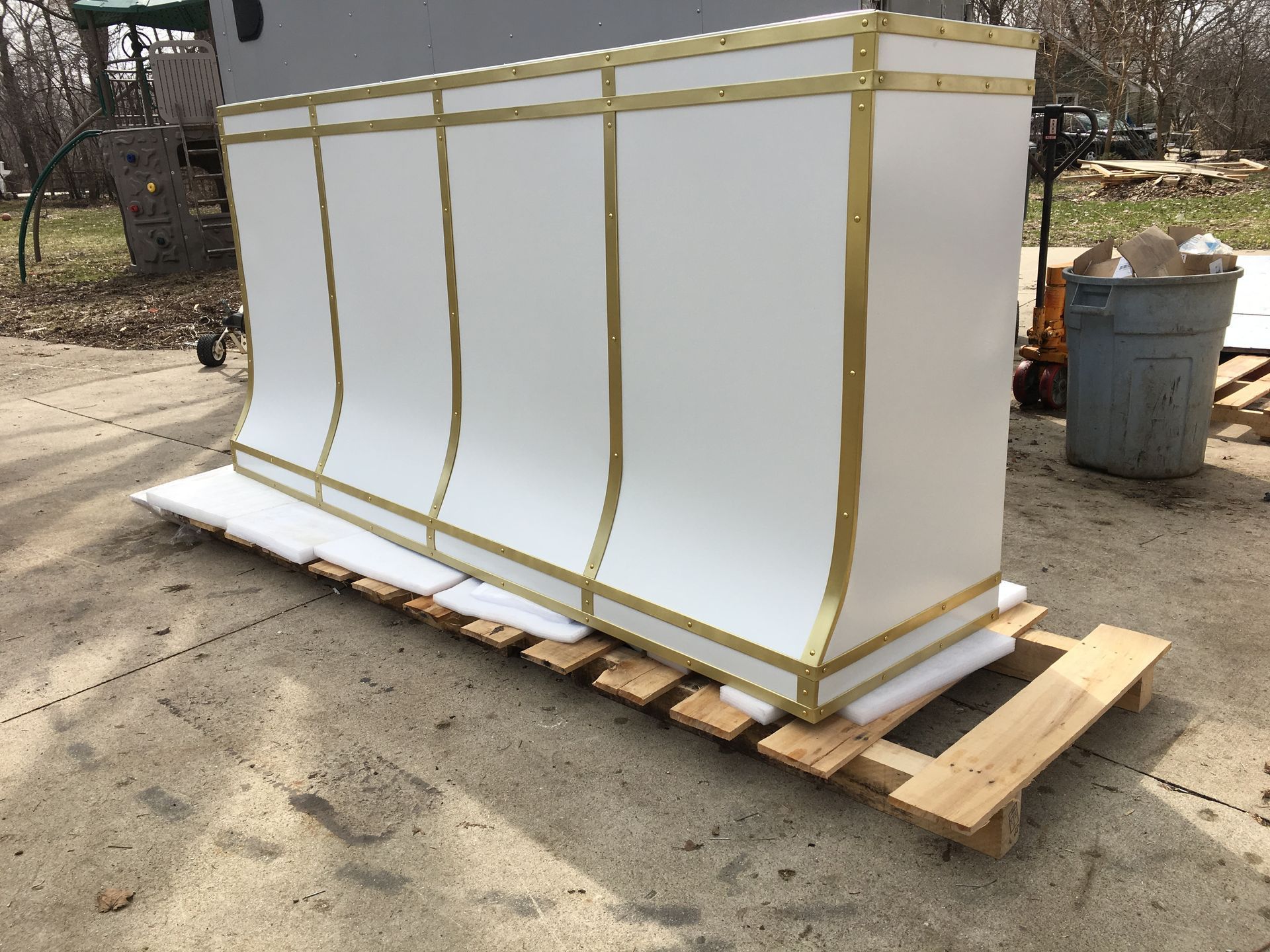 White rectangular equipment crate wrapped in gold straps, sitting on a wooden pallet outdoors.