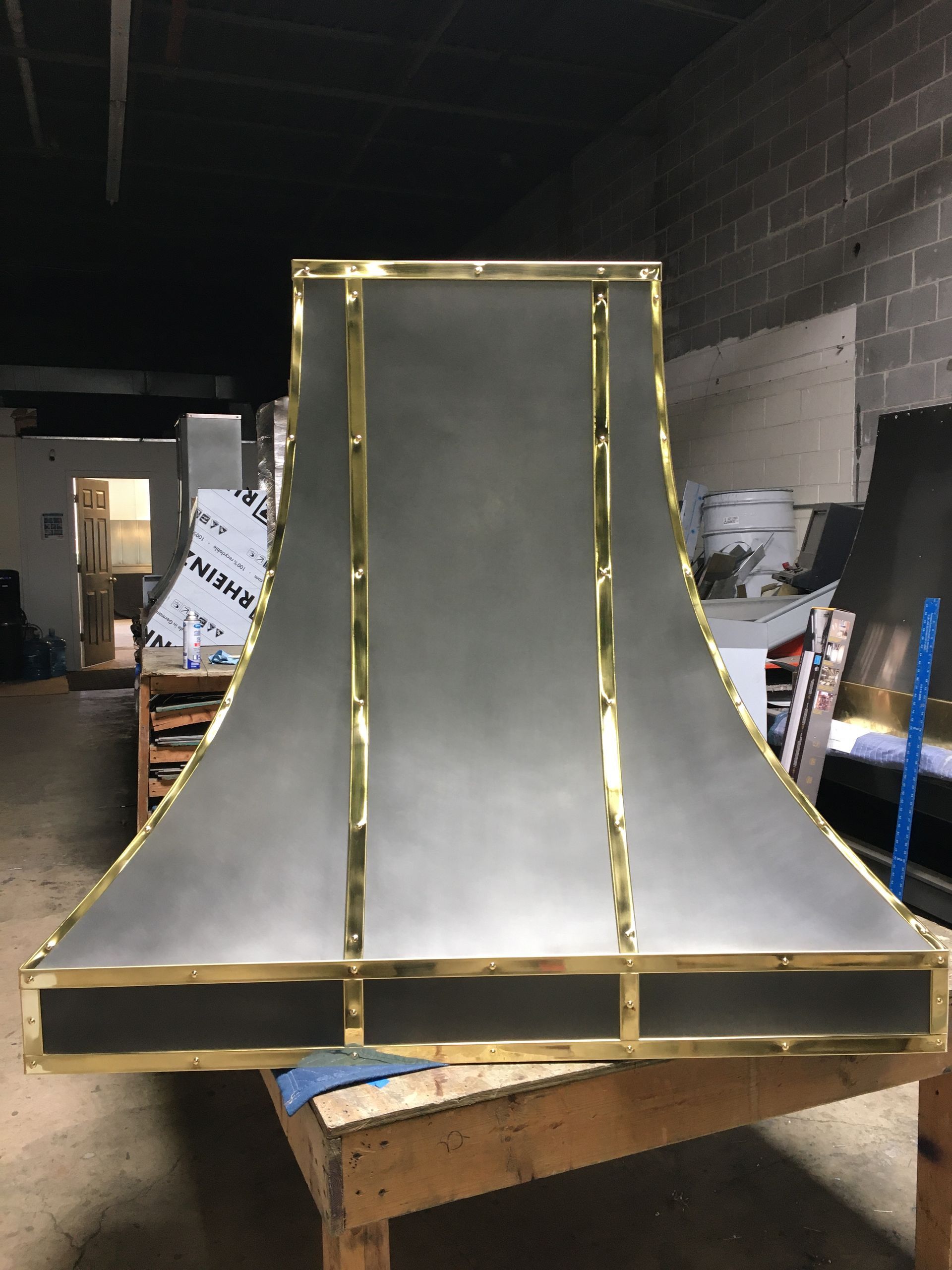 Metal duct hood with gold trim standing on a workbench in a warehouse workshop
