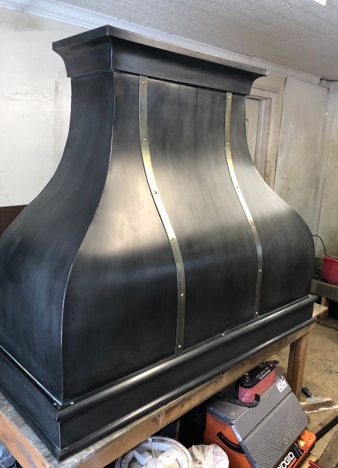 Black metal range hood with curved silver panels in a workshop or kitchen setting